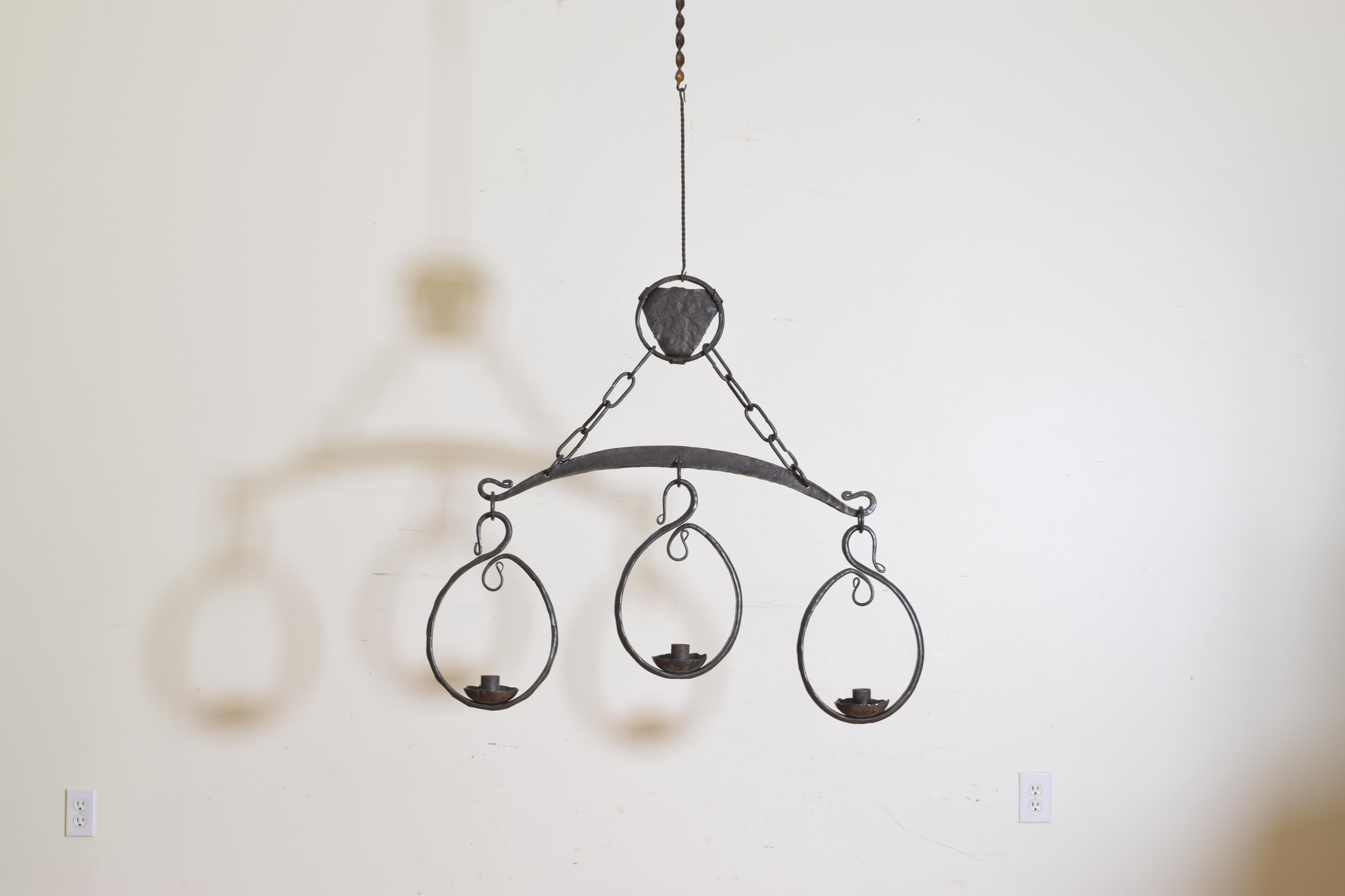 Hanging from a shaped metal and iron medallion this chandelier has a bow shaped body with three hanging ovals, each with a centered bobeche.