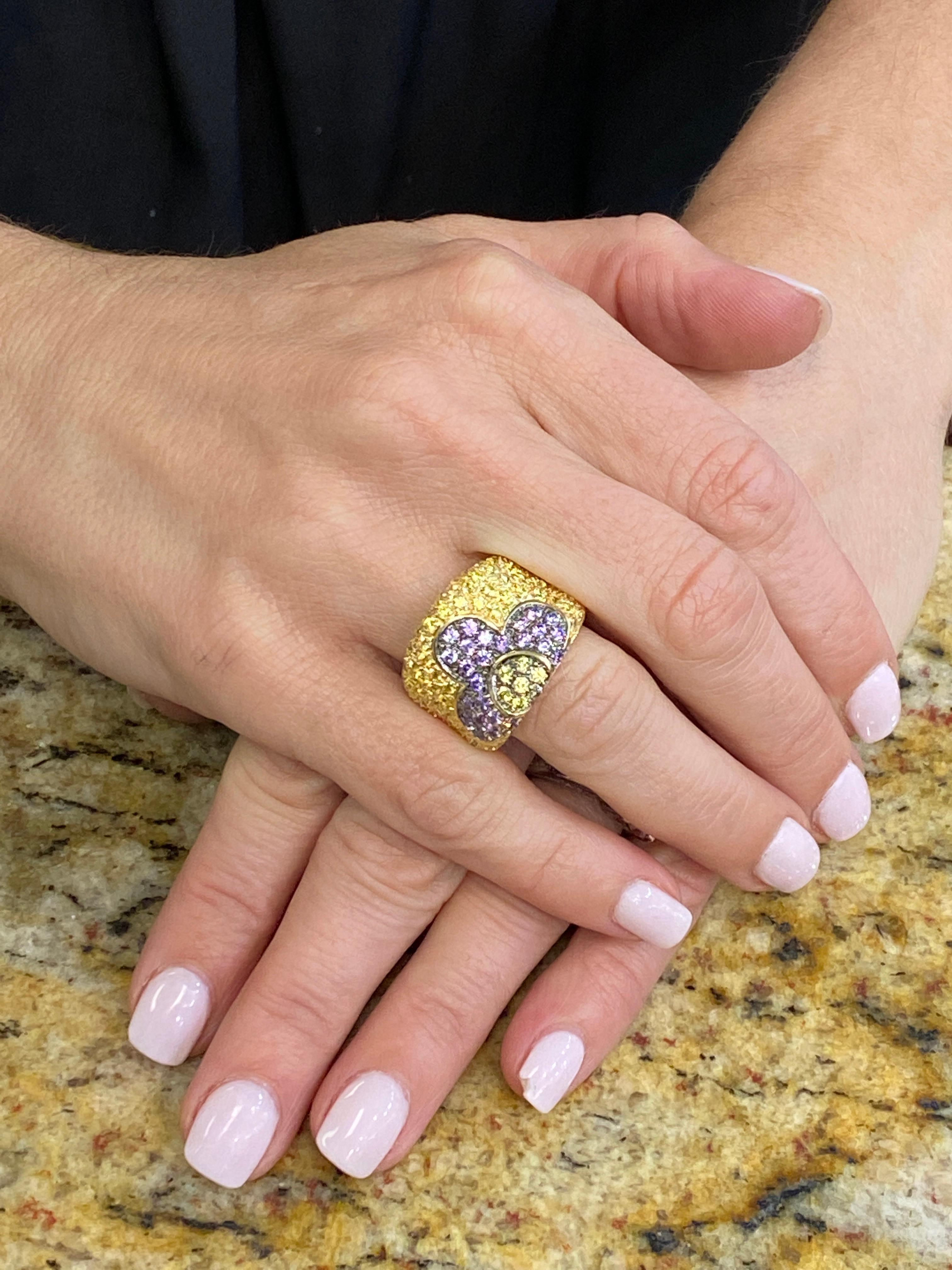 Italian yellow and purple sapphire floral design band fashioned in 18 karat yellow gold. The ring features round brilliant cut yellow and purple sapphire gemstones in a floral design. The ring measures .75 inches in width and is currently size 7.0