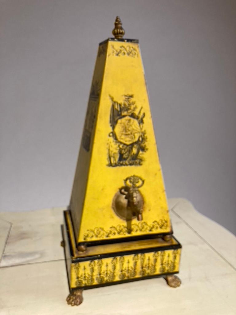 Eine bezaubernde neoklassizistische gelbe Tole-Wasser- oder Tee-Urne in Form eines Obelisken, der auf einem quadratischen Sockel mit vier Kugeln balanciert, die von Messingfüßen getragen werden. Dekoriert auf vier Seiten mit Karten der Regionen von