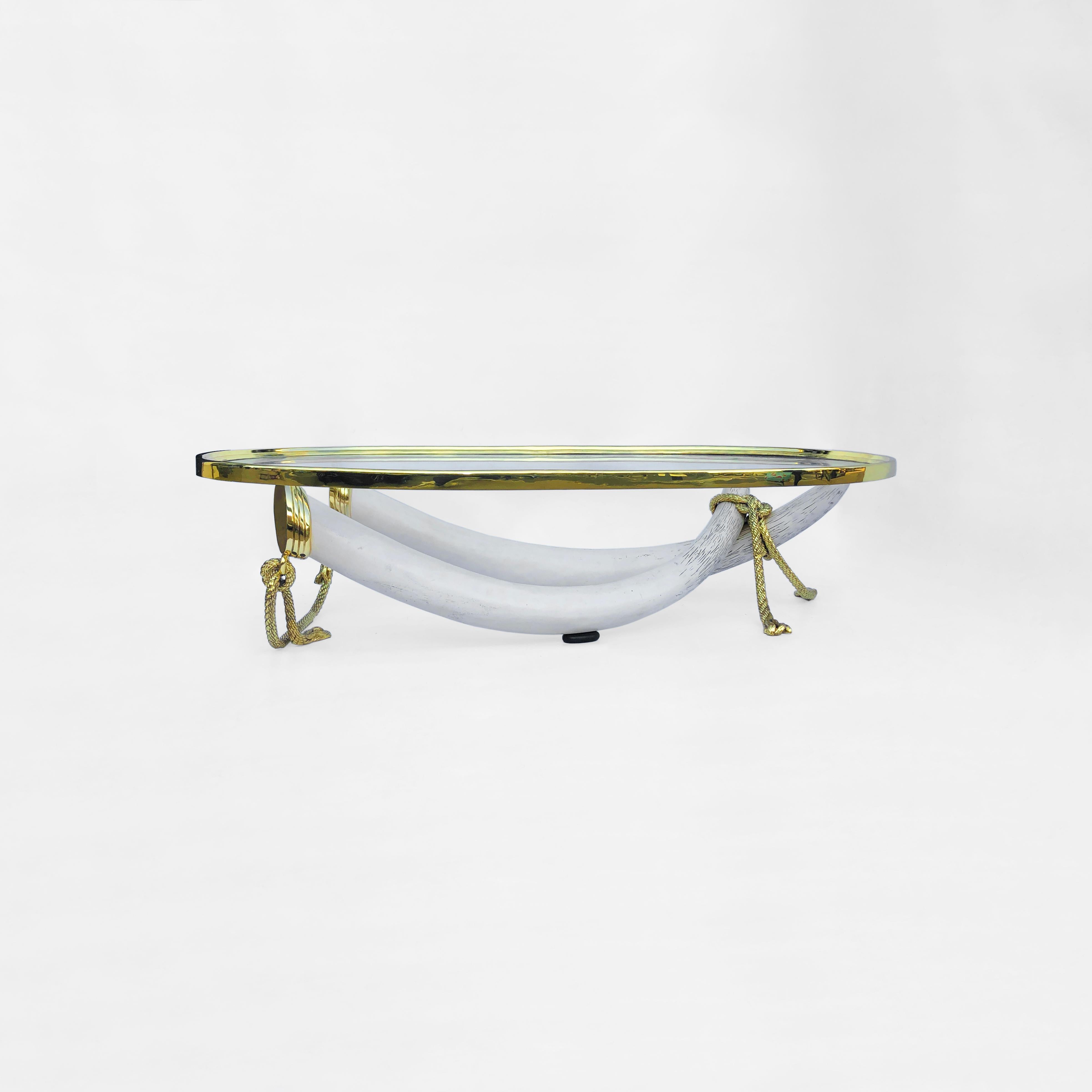 Dieser massive Couchtisch der spanischen Designer Valenti ist eines der beeindruckendsten Stücke, die wir seit langem gesehen haben. Er besteht aus zwei gekreuzten Stoßzähnen aus Elfenbeinimitat, die eine große, ovale Platte aus Messing und Glas
