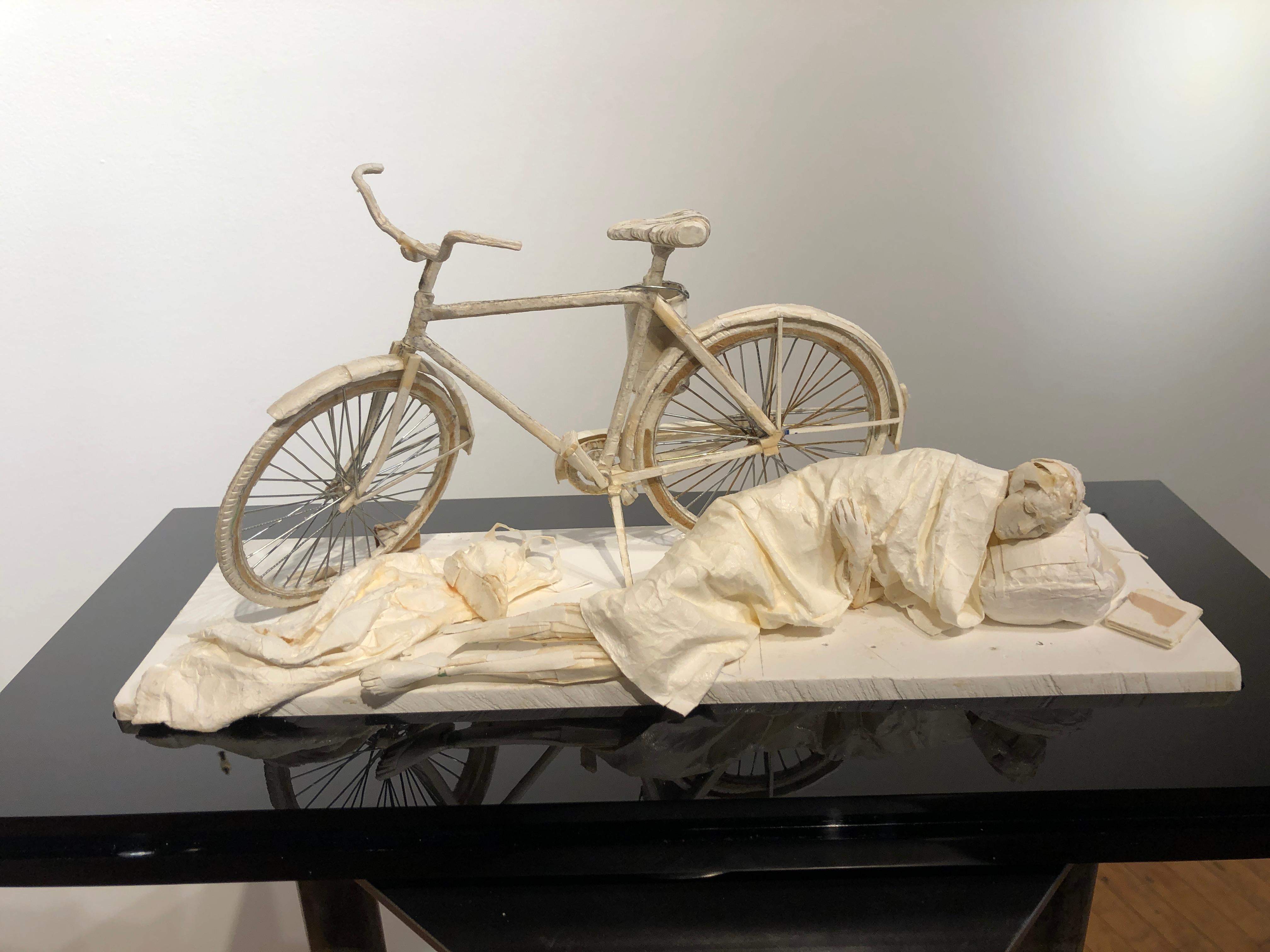 Cette sculpture réaliste est créée à partir de bouts de papier et de colle.  La sculpture s'inspire des personnages que l'on trouve dans les villes du monde entier.  Dans ce cas, un personnage dort à côté d'un vélo.  Un sac à dos est utilisé comme