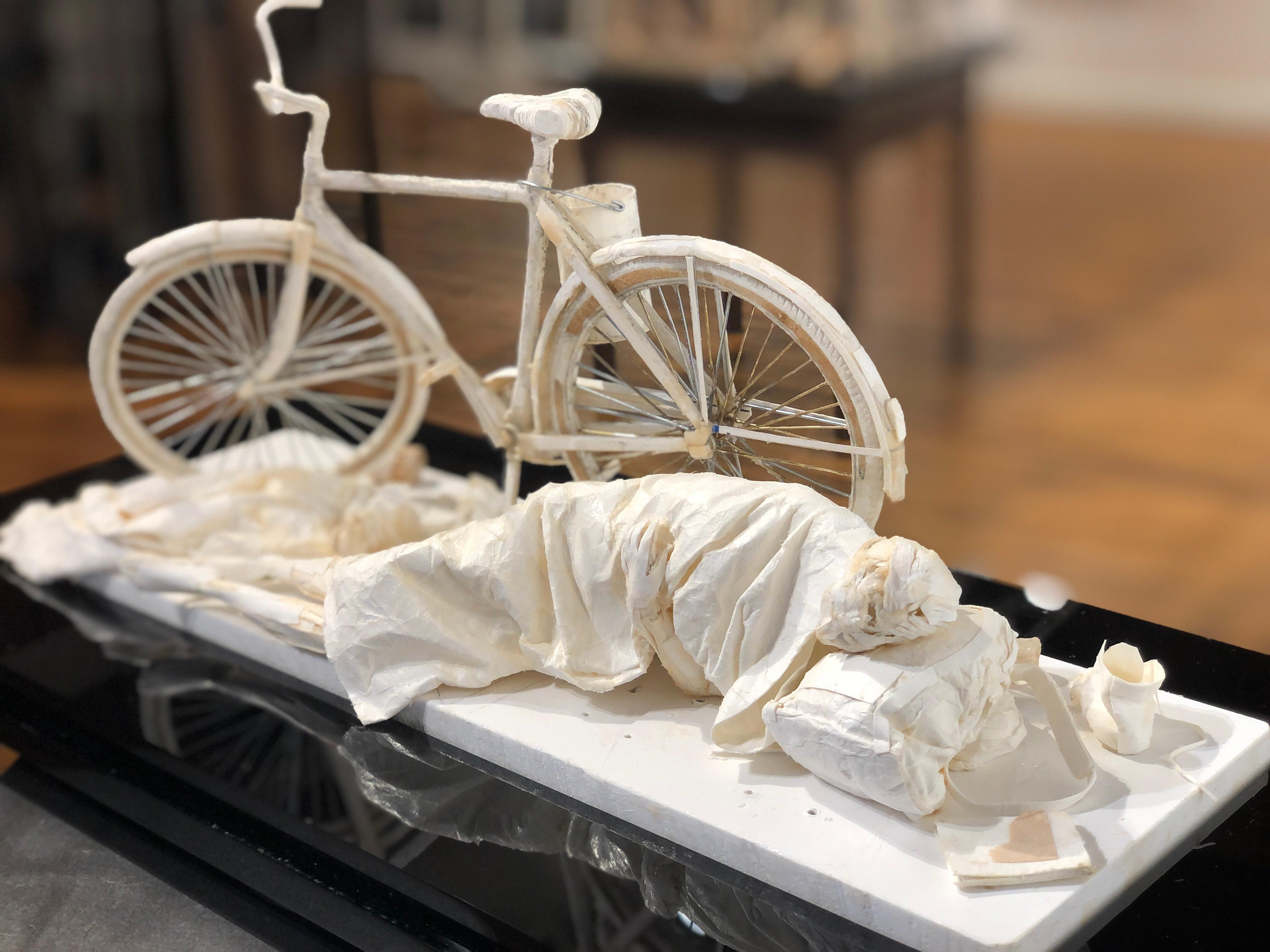 This realistic sculpture is created from scraps of paper and glue.  The sculpture is inspired by the characters found in cities around the world.  In this case, a figure sleeps beside a bicycle.  A backpack is used as a pillow and shoes has been