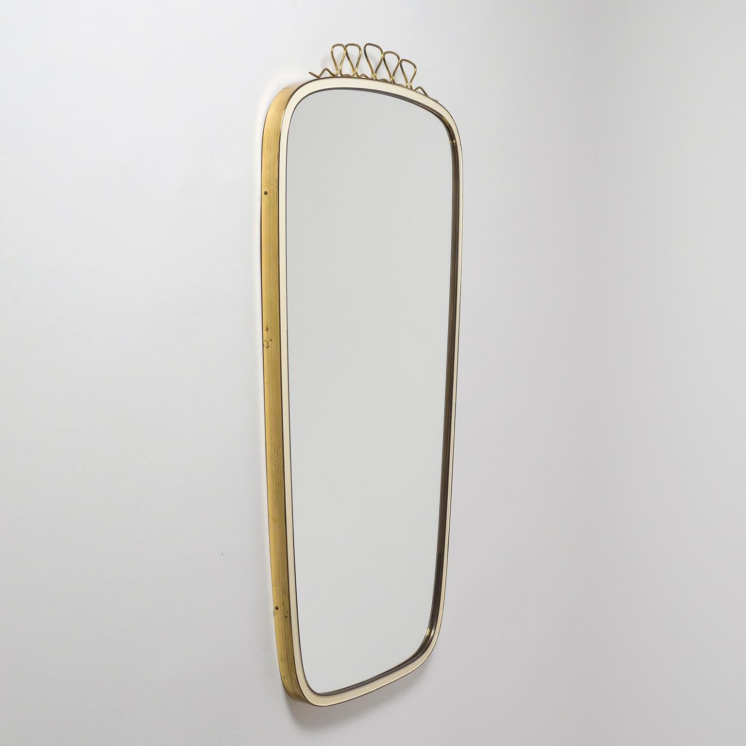 Lovely midcentury mirror with a sculpted brass rim which has a light ivory enamel coating on the inlet. Topped off with a charming undulating brass finial. Very good original condition with a bit of patina on the brass. Measures: Mirror size is