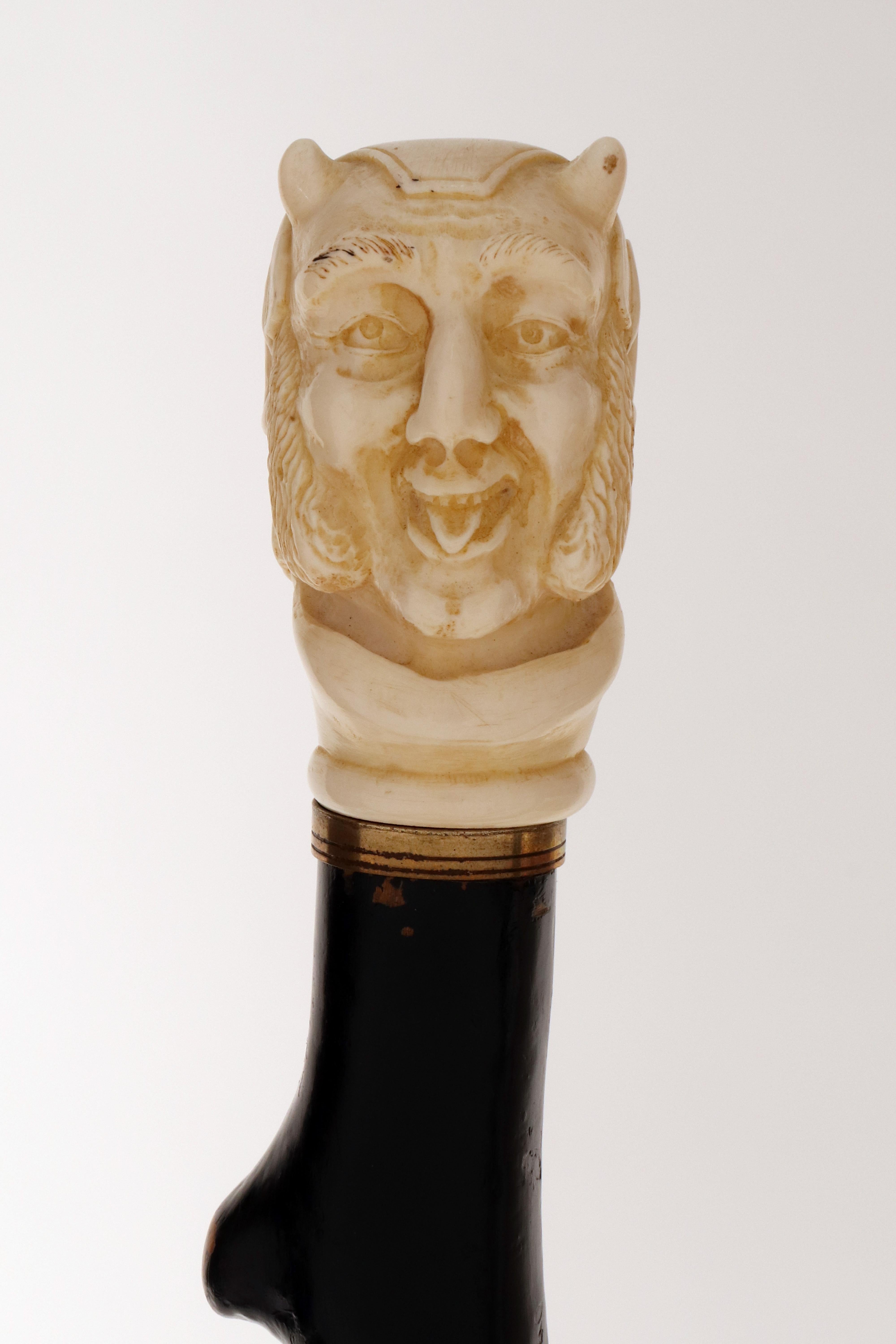 Bastón: mango tallado en marfil. El mango representa un rostro caricaturesco de Mefistófeles. Asta de madera de frutal ebonizada de forma rústica. Anillo de plata. Virola metálica. Inglaterra, hacia 1870. (SÓLO ENVÍO A LA UE)