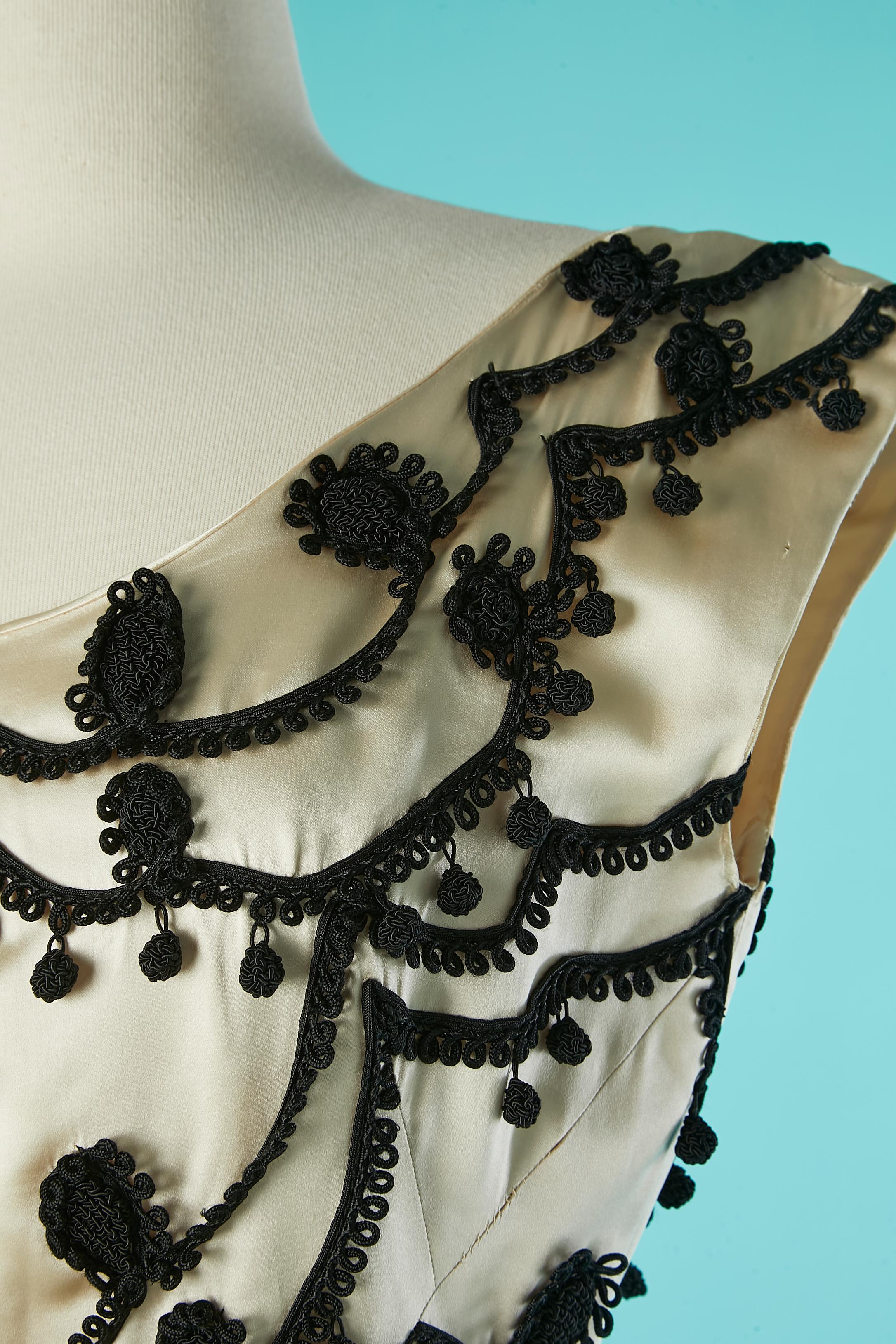 Robe de cocktail en satin ivoire avec passementerie noire. Pas d'étiquette de composition du tissu mais probablement une doublure en organza sur la partie supérieure de la robe et une doublure en acétate à l'intérieur de la jupe. Gros grain