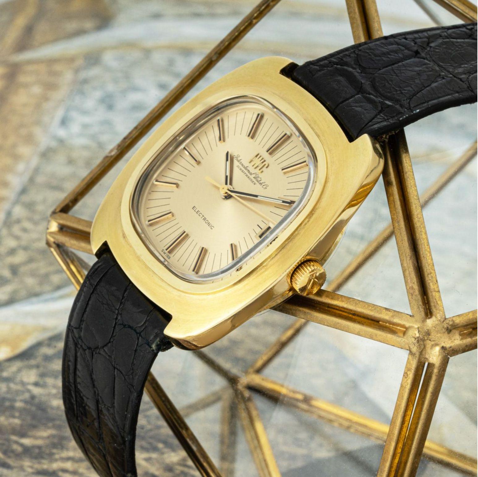 A mens yellow gold IWC Electronic Old Tuning Fork wristwatch. Featuring a champagne dial with applied hour markers, a date aperture and a yellow gold bezel. Fitted with a plastic glass, a manual winding movement and an original black leather strap