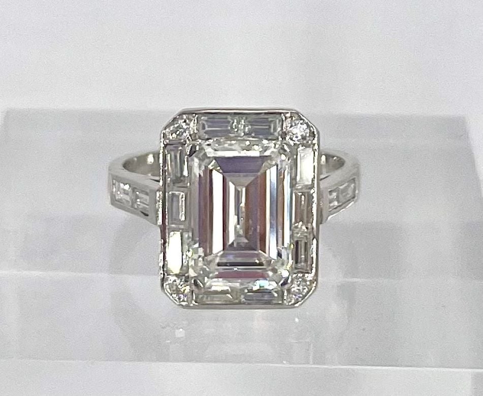 This unique Art-Deco inspired engagement ring celebrates the clean lines and glamour of the era. The ring features a 2.60 carat emerald cut diamond, certified by GIA to be F color and SI2 clarity. The diamond is surrounded by a sleek halo of