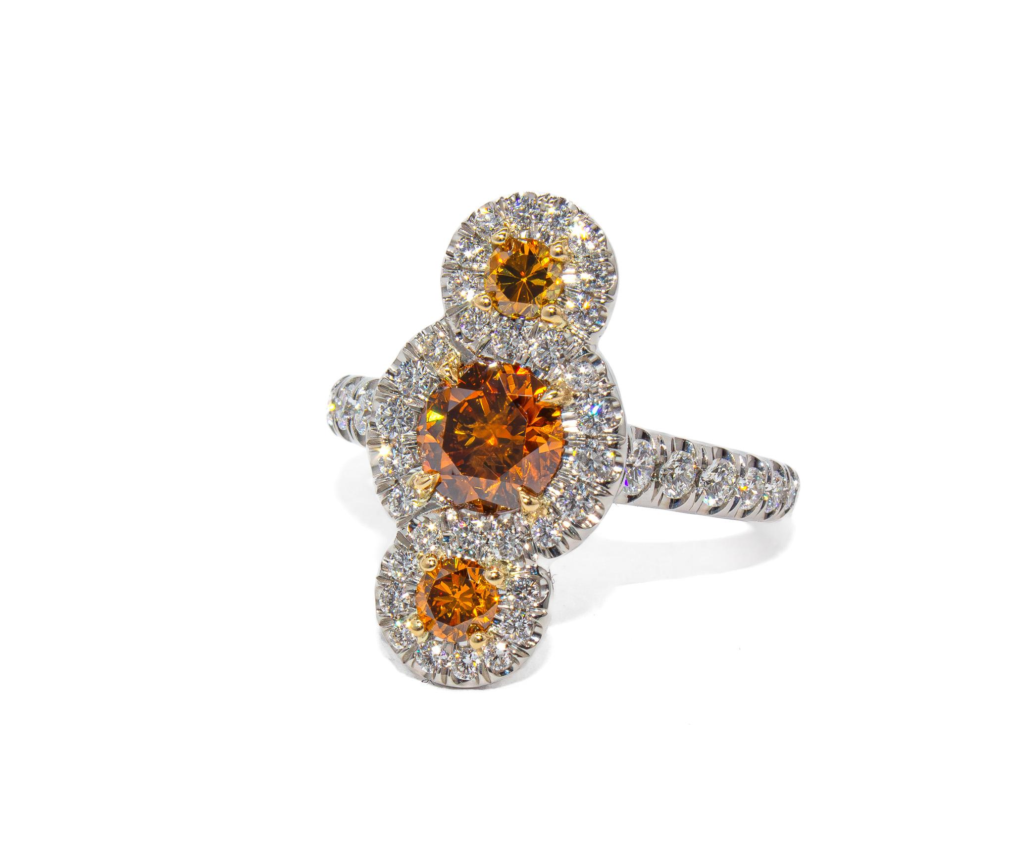 This one-of-a-kind three-stone fashion ring from J. Birnbach is a striking study in warmth and contrast. The center diamond is a 0.90 carat round brilliant, certified by GIA as Fancy Deep Brownish-Orange—a vivid and richly saturated hue that feels