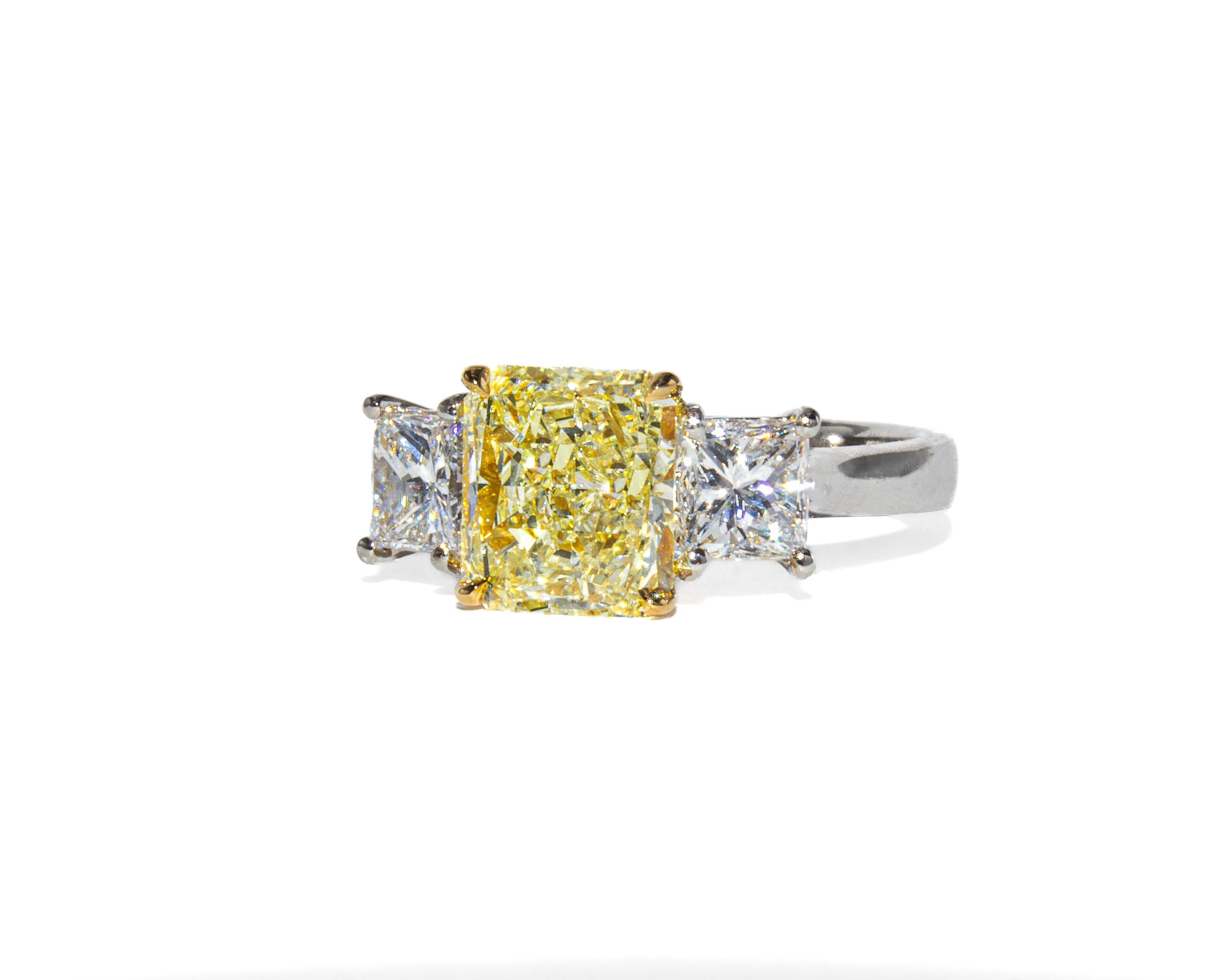 Una sorprendente mezcla de color y artesanía, este anillo de tres piedras hecho a mano incluye un diamante de talla radiante de 1,76 quilates, certificado por GIA como Amarillo Claro Fancy con claridad SI1. La vibrante piedra central está flanqueada