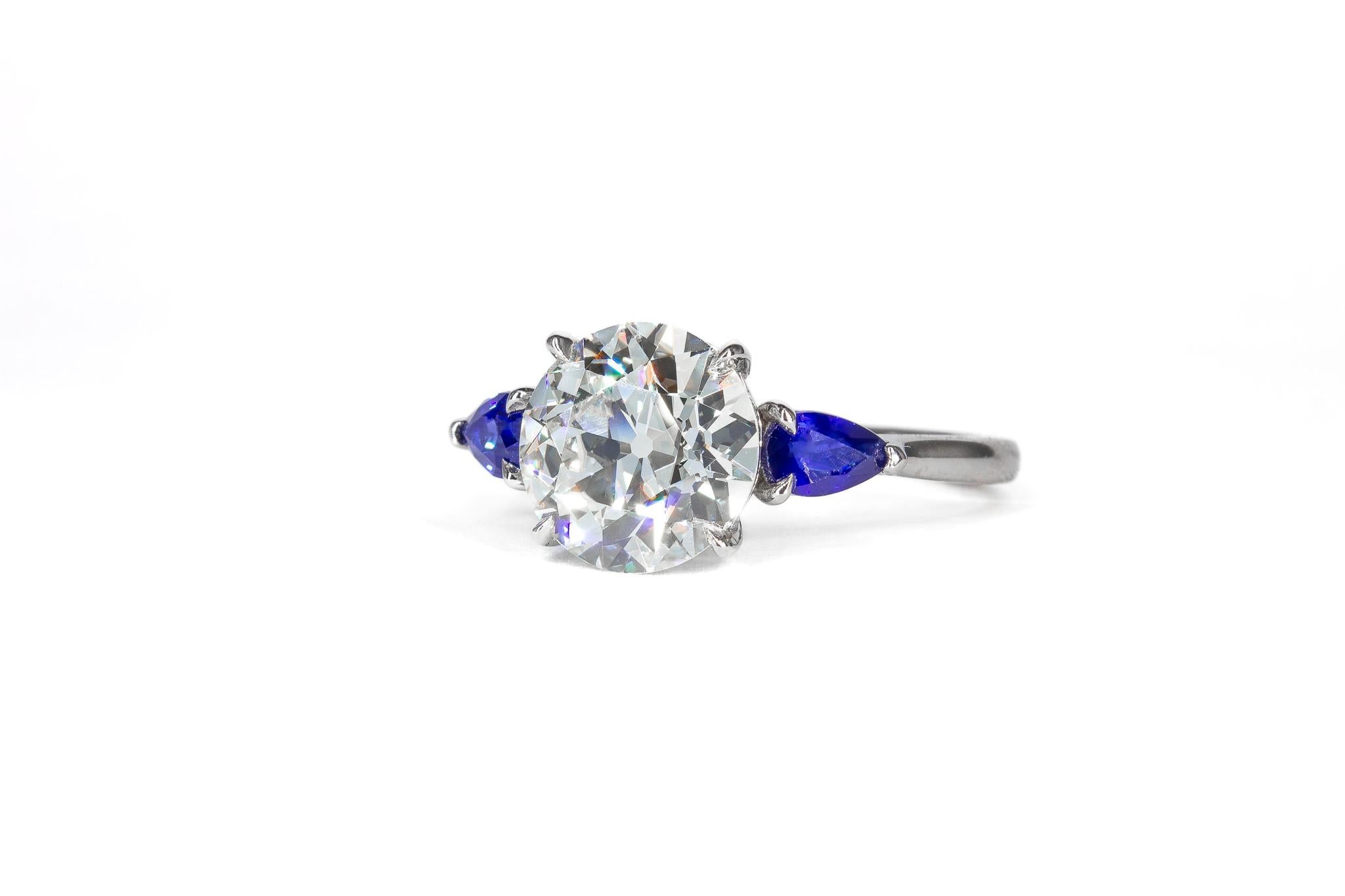 This exquisite platinum three-stone ring showcases a captivating 2.51 carat old European cut diamond, certified by GIA as N color and VS1 clarity. The center stone’s vintage charm is complemented by two pear-shaped sapphire side stones, totaling