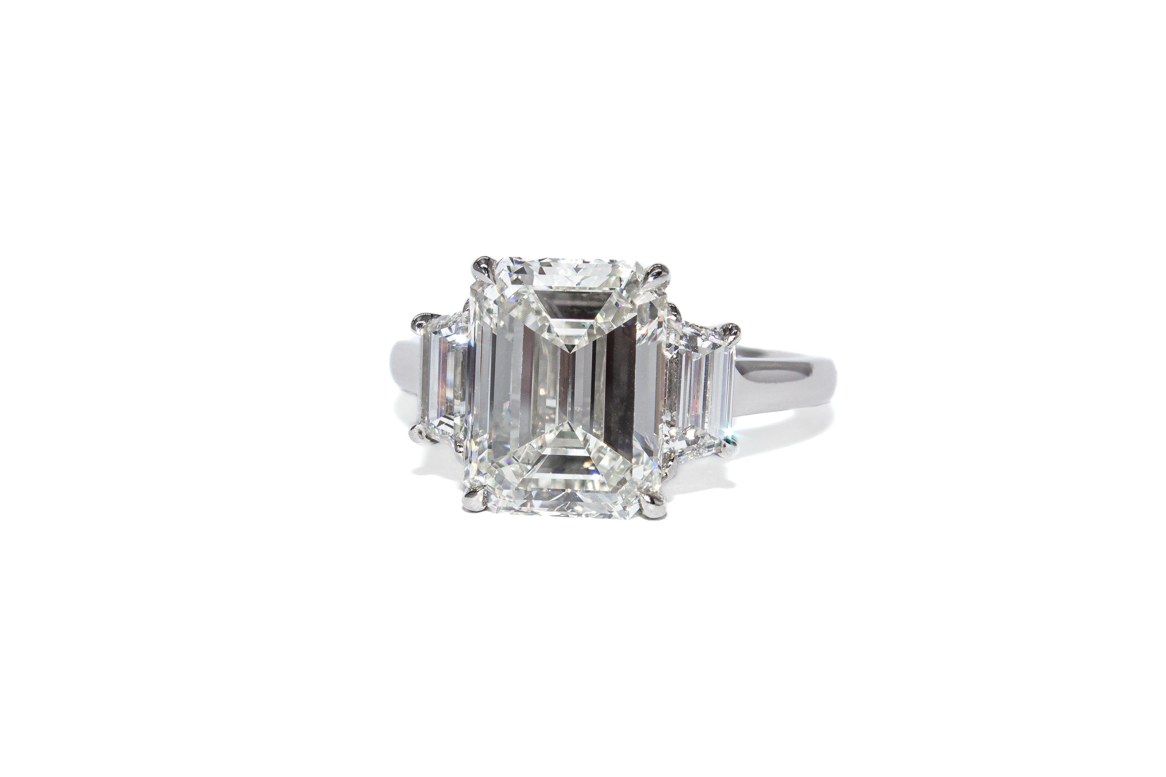 Cette élégante bague en platine à trois pierres met en valeur un diamant certifié de taille émeraude de 4,01 carats, de couleur J et de pureté VS2, selon la description du GIA. Les facettes allongées de la pierre centrale, taillées en escalier,