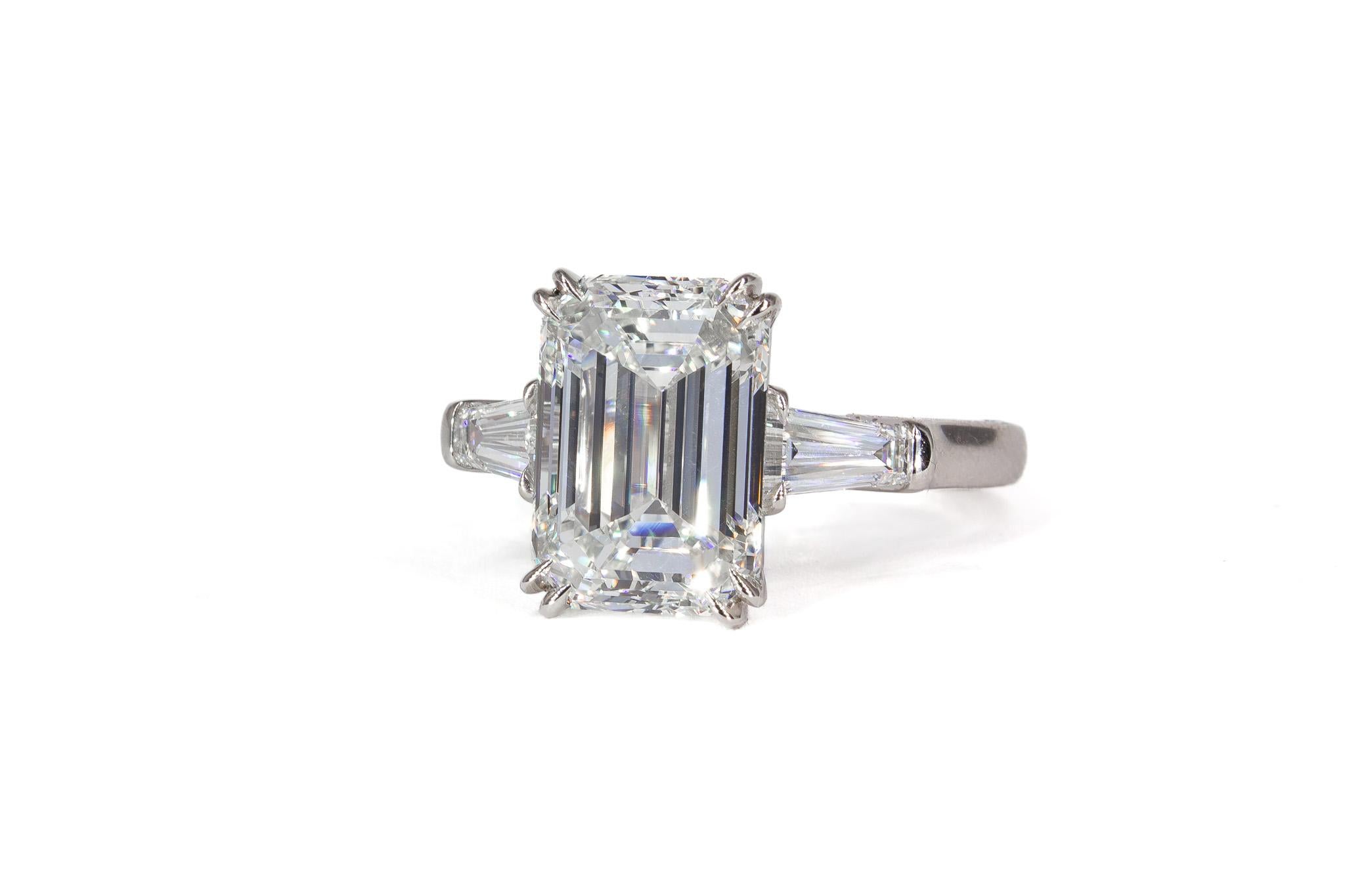 This timeless three-stone ring is a stunning celebration of classic elegance and exceptional craftsmanship. At its center is a GIA-certified 5.03 carat emerald-cut diamond with a captivating K color and VS2 clarity. The sleek emerald cut showcases