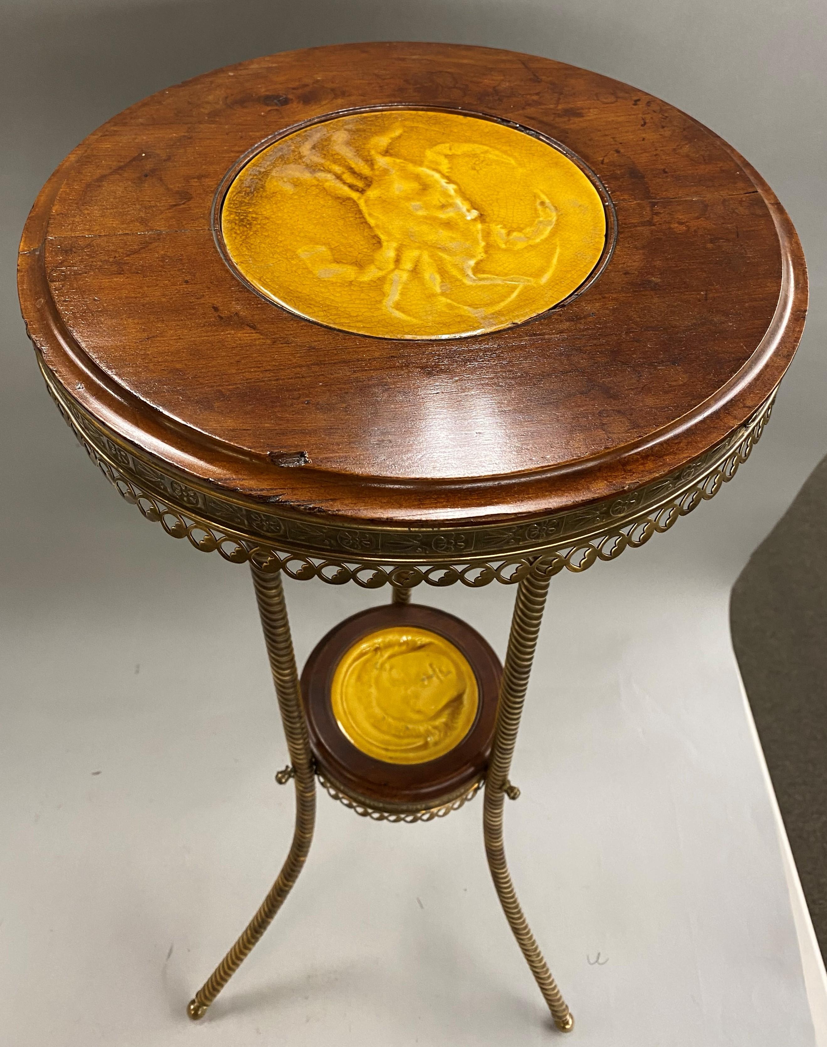 A beautiful round wooden and brass pedestal or tripod stand with butterscotch tile inserts in the top and medial stretcher, the upper tile featuring a crab, signed on tile base “J. & J.G. Low, Patent, Art Tile Works, Chelsea, Mass, USA, copyright