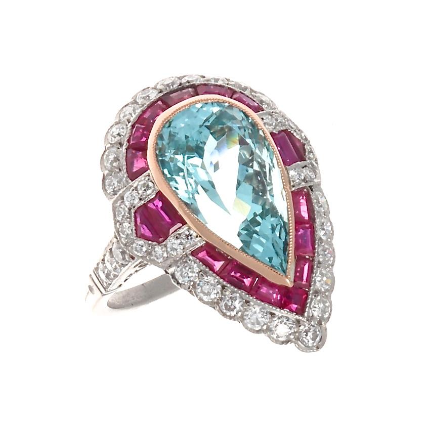 A symphony of color and geometric balance that is exemplary of the fantastically stylish art deco era. Featuring a glowing 6 carat pear shaped aquamarine surrounded by expertly calibrated vibrant red rubies and perfectly complimented by colorless