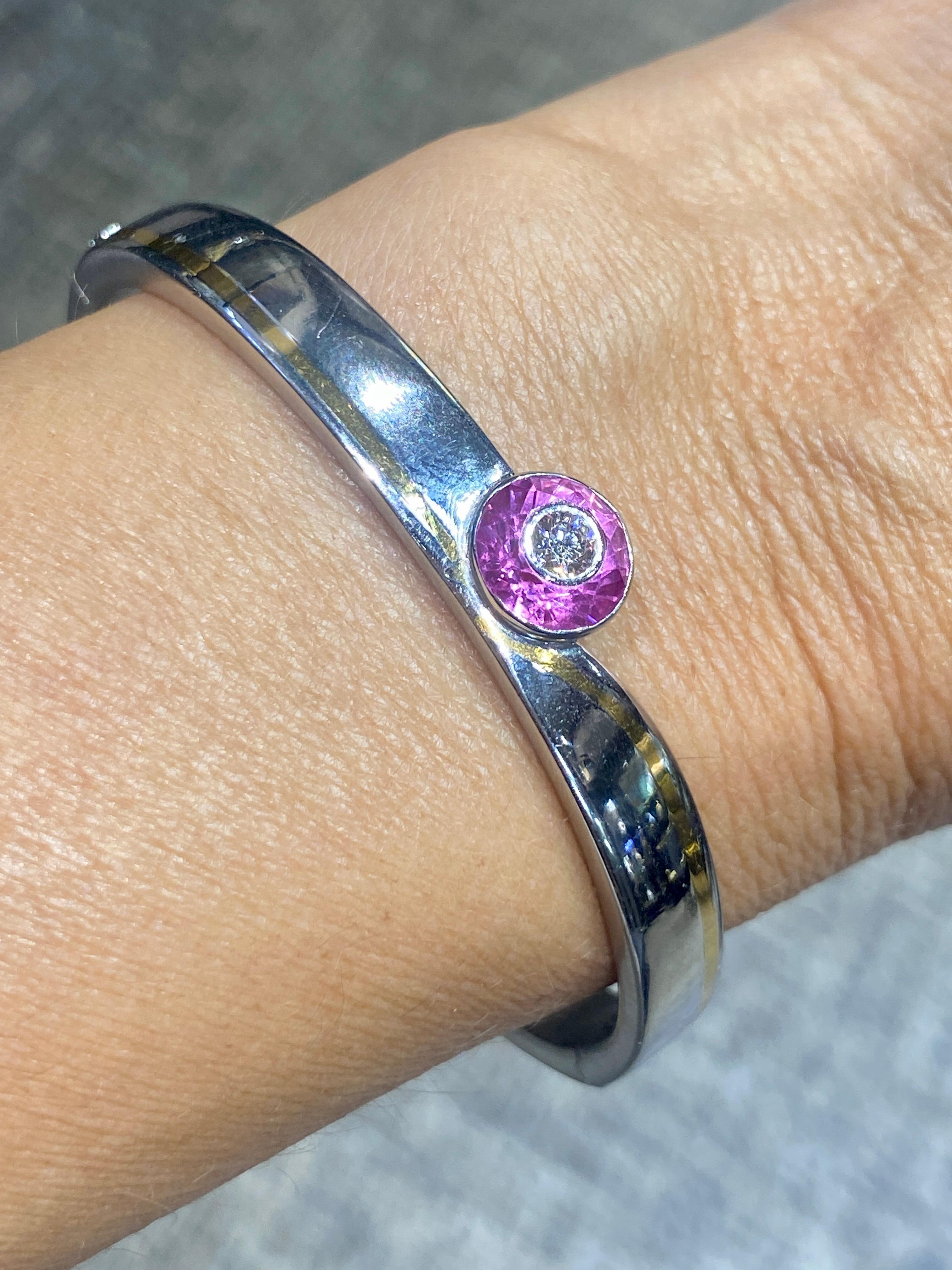 This modern Jacob & Co platinum bangle's design plays with the polished and matt aspects of the metal. The central stone is a beautiful round cut pink sapphire that has been carved to hold the round cut diamond. A strand of yellow gold runs along