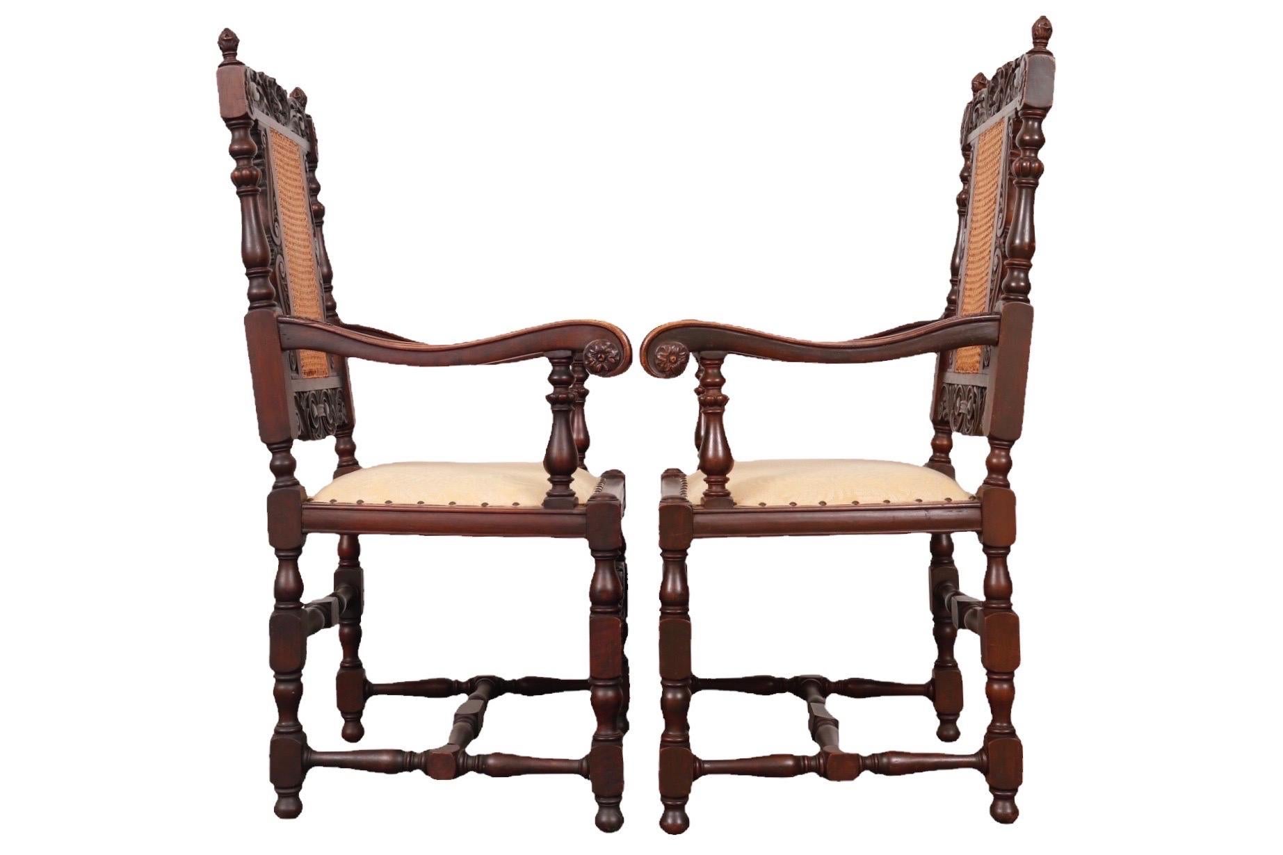 A pair of ornately carved Jacobean style armchairs. Back splats are double caned and topped with scrolled acanthus leaves each side of a central scallop shell. Square seats are upholstered with a light yellow fern damask secured with nailheads.