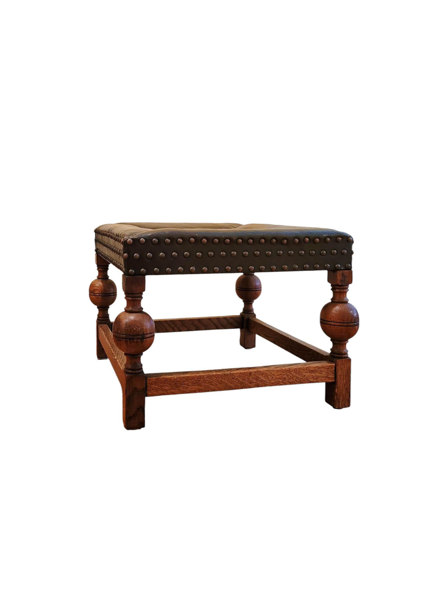 A square Jacobean Revival footstool, raised on boldly turned bobbin legs with a sturdy box stretcher base. The seat is upholstered in dark leather with stitched detailing, while the apron is finished with three rows of decorative nailheads. Likely