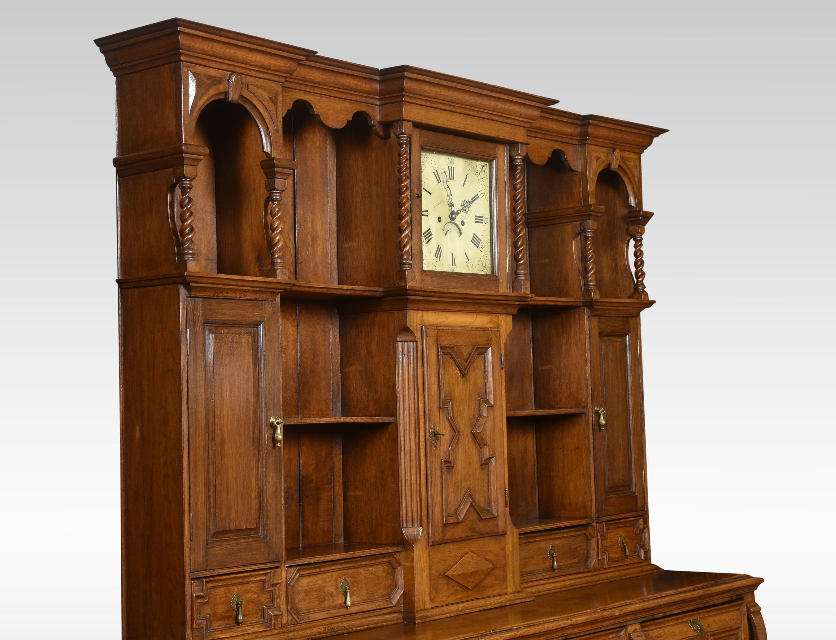 Jacobean style oak dresser, having a deep boarded rack with canopy frieze above an arrangement of shelves, drawers and cupboards, surrounding the eight-day clock with brass dial signed Torkington Newcastle, striking on a bell. The base section is