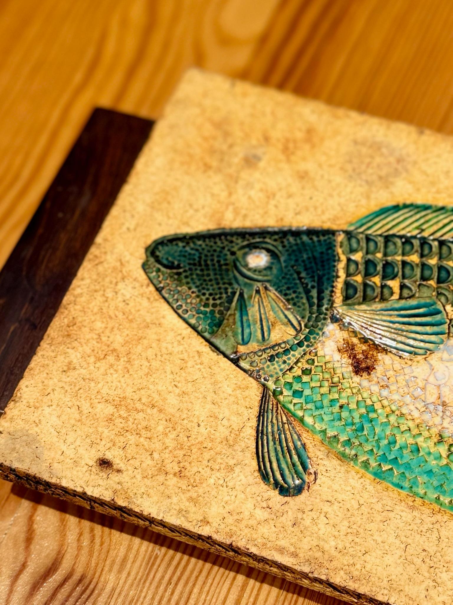 A captivating rectangular ceramic plaque depicting a stylized fish in raised relief, intricately detailed with textured scales and glazed in a harmonious palette of turquoise, green, and cream. The rich, layered glazes create subtle variations in