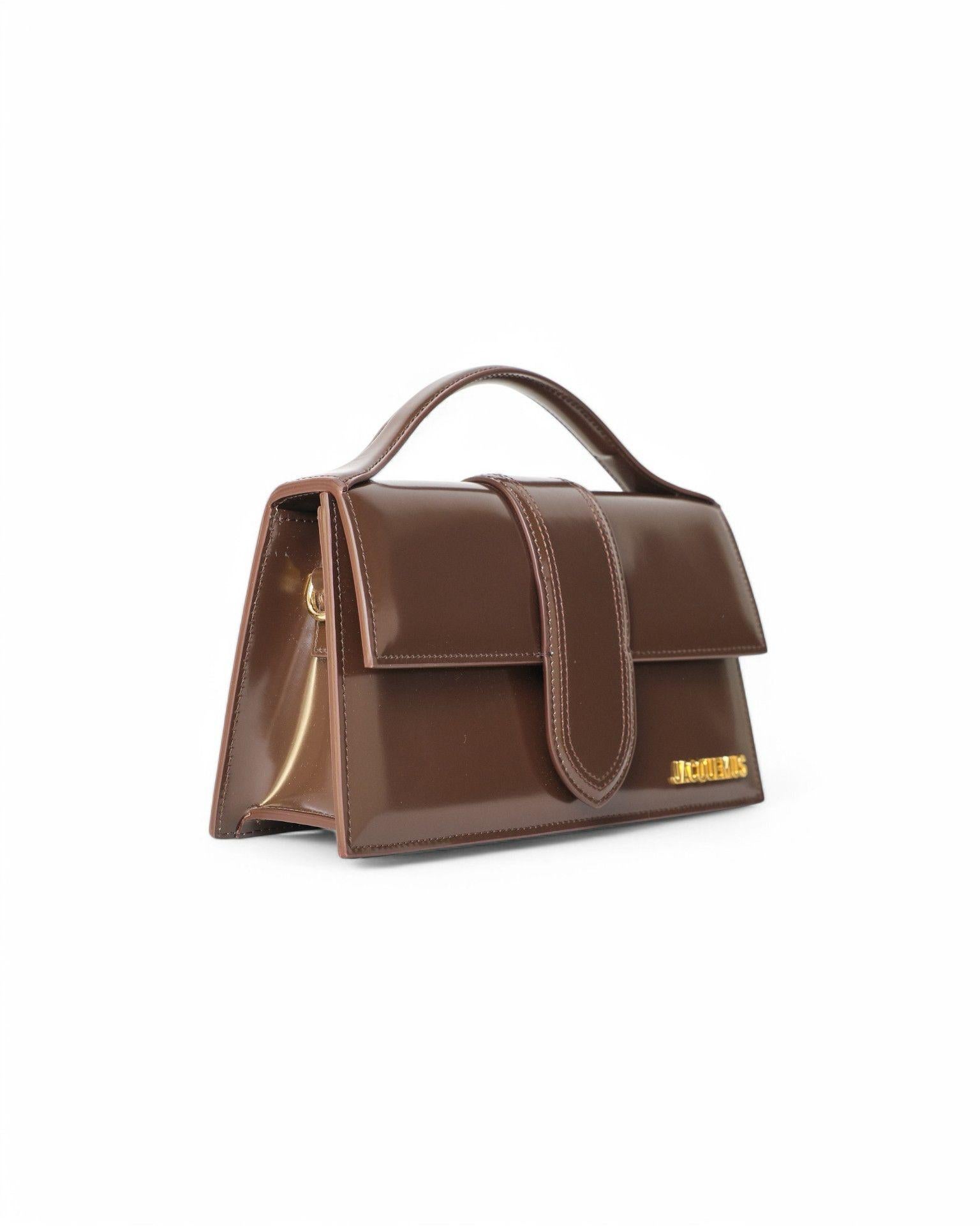 Jacquemus signature bag, model Le Grand Bambino, made of shiny leather in brown color with gold hardware. Featuring a front flap with magnetic button closure, internally lined with beige canvas, roomy for essentials. Munita di un manico rigido