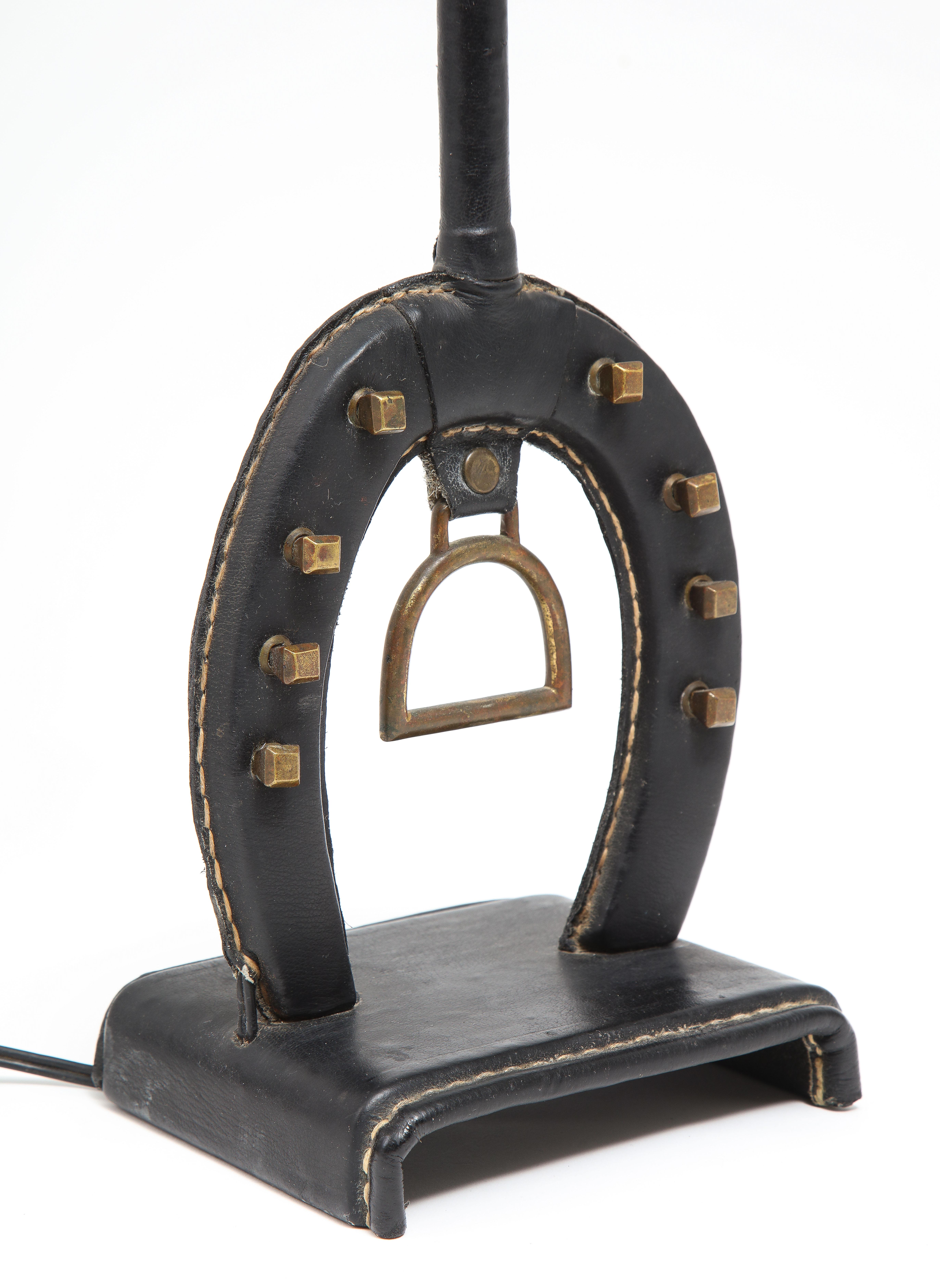 Jacques Adnet lamp with Equestrian theme, the body of the lamp is a horseshoe with its distinctive nail pattern in brass; it rests on a bent metal base, wrapped in Adnet distinctive stitched leather. Base only is 16x5x6. Shade is for photos only,