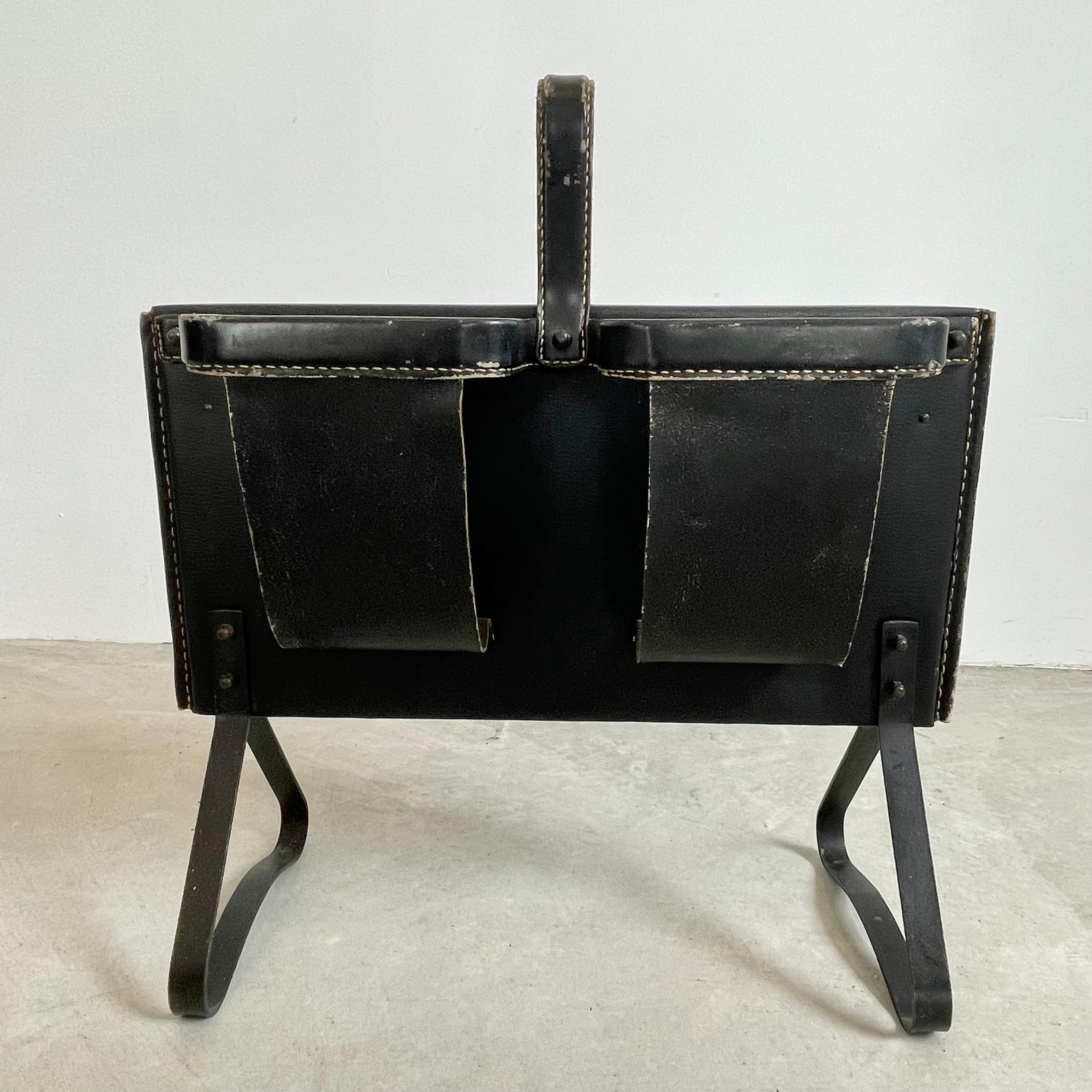 Handsome leather and iron magazine rack by French designer Jacques Adnet. Black leather frame with signature Adnet contrast stitching. Leather loop handle at the top. Two compartments on one side, and one larger compartment on the opposite side.