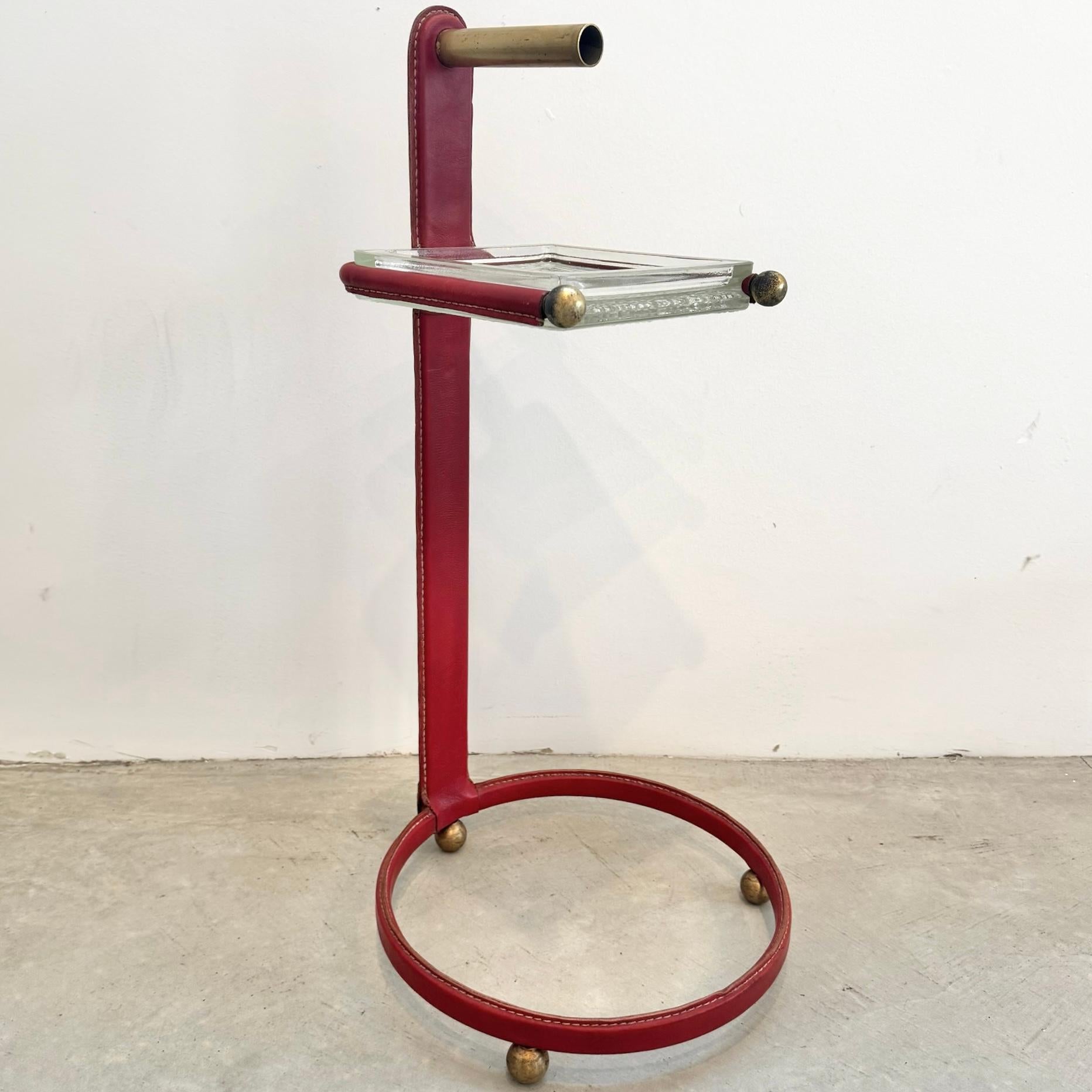 Stunning red leather side table / catchall  by French designer Jacques Adnet. Iron frame wrapped in a beautiful red leather with glass catchall on top. A large ring with three brass ball feet make up the base. It features contrast stitching along