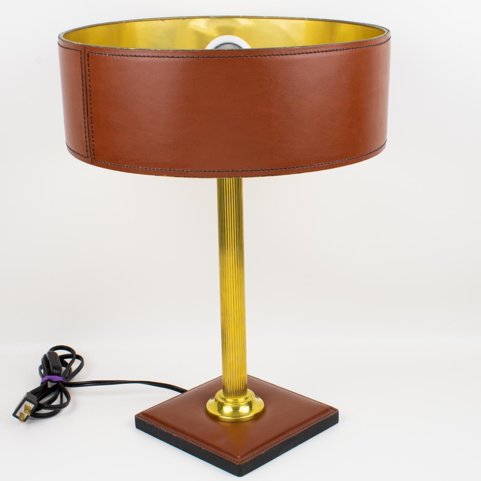 This elegant table lamp was designed by Jacques Adnet (1901 - 1984). The classical cinnamon brown color lamp features a hand-stitched shade with gilded paper lining, polished brass reeded column stem, and a square base in the same hand-stitched