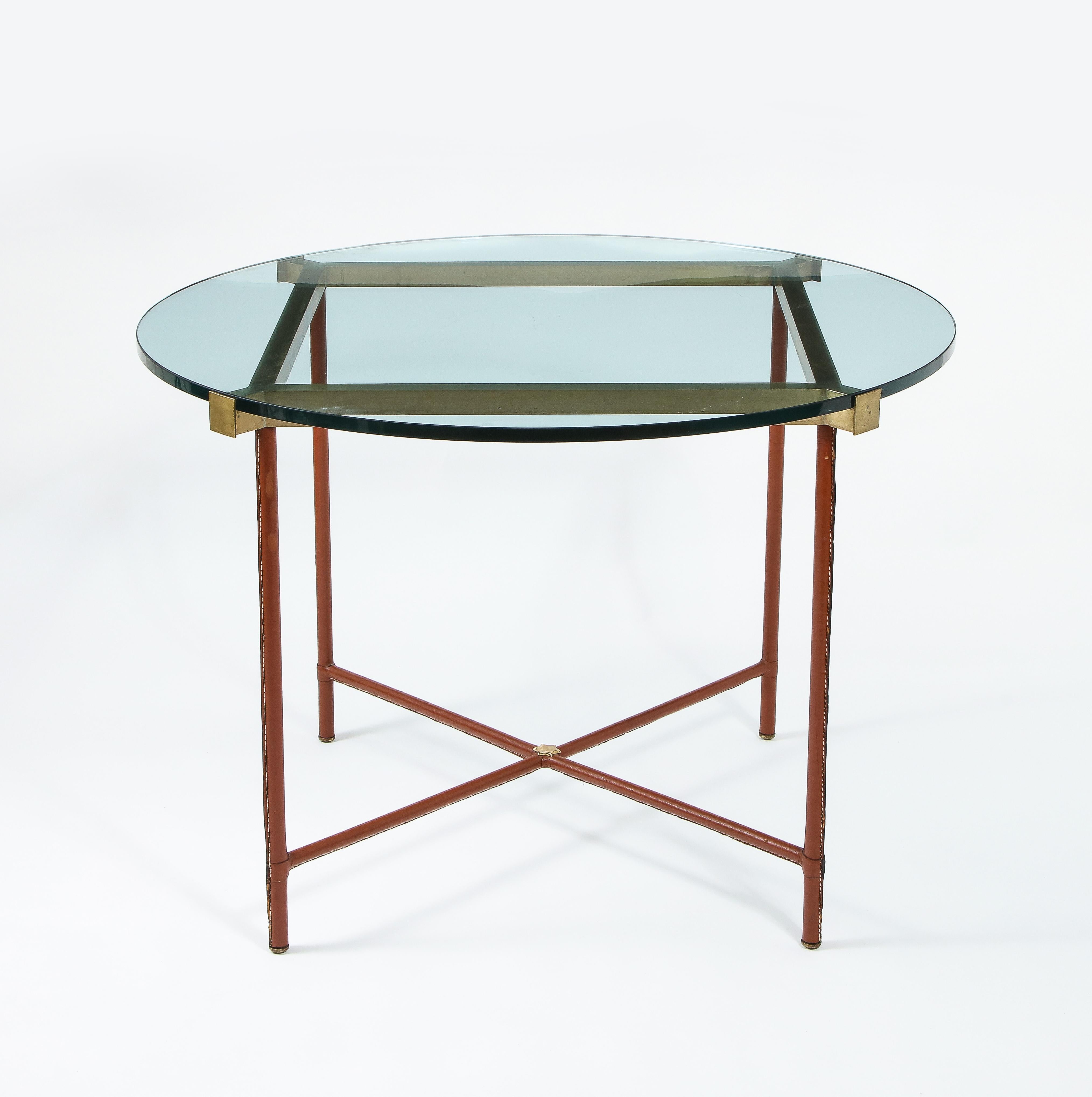 Exceptionnelle table centrale de Jacques Adnet habillée de cuir brun tabac, ses quatre pieds réunis par un X et un large cadre en laiton supportent le plateau en verre, elle repose sur des pieds en bronze.