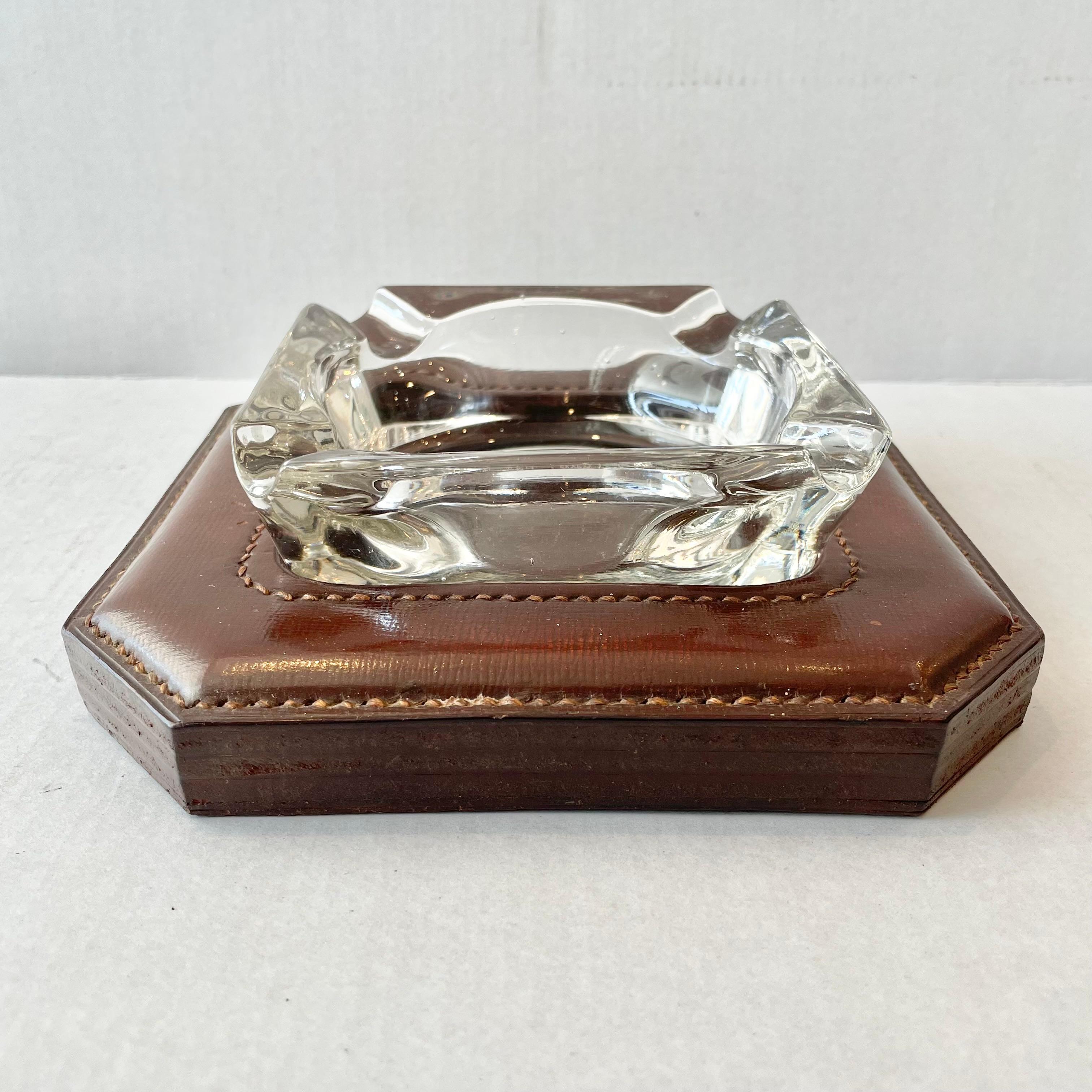 Classic leather and glass catchall / ashtray by French designer Jacques Adnet. Rich cognac leather base with signature Adnet contrast stitching and a circular inset cutout. Crystal glass with circular base sits nicely inside with four cigarette