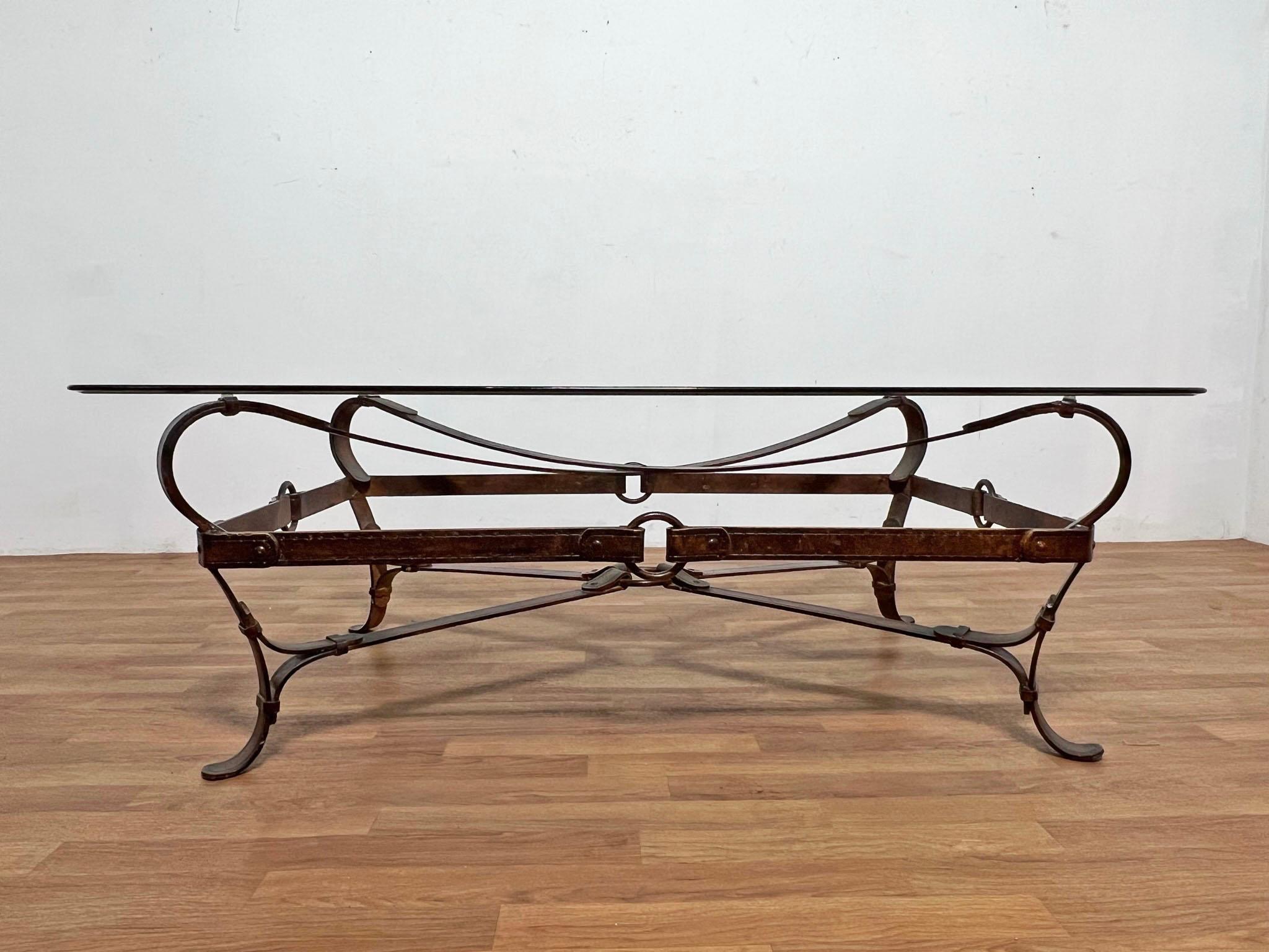 A cocktail table featuring a trompe l’oeil framework of equestrian harnesses rendered in wrought iron, in the manner of Jacques Adnet for Hermes, circa 20th century. Glass top with rounded corners and beveled ogee edge.