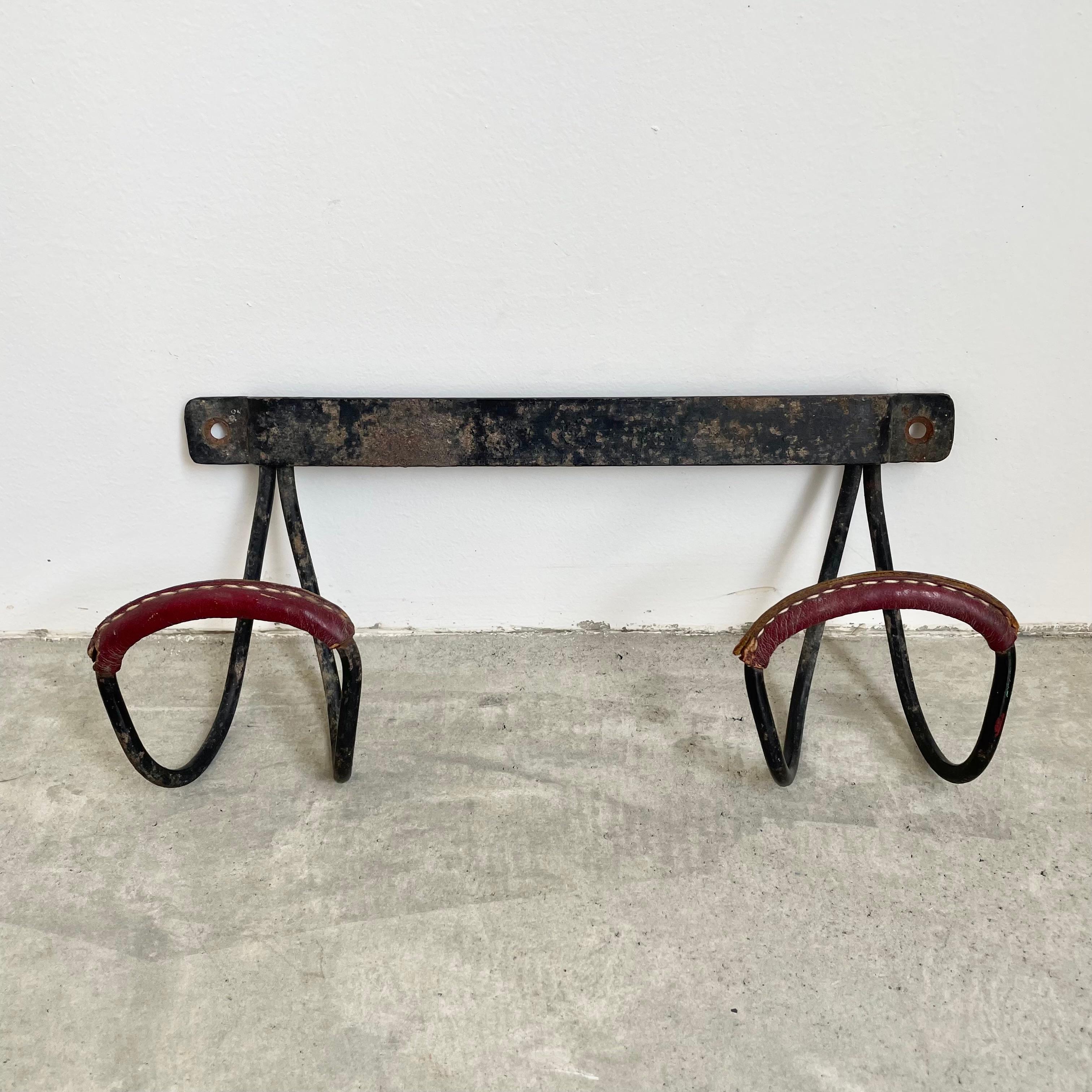 Handsome wall rack by French designer Jacques Adnet. Frame made of solid iron and features two large tongue hooks with red oxblood leather detailing on each hook. Signature Adnet contrast stitching. Good vintage condition. Priced individually. 

