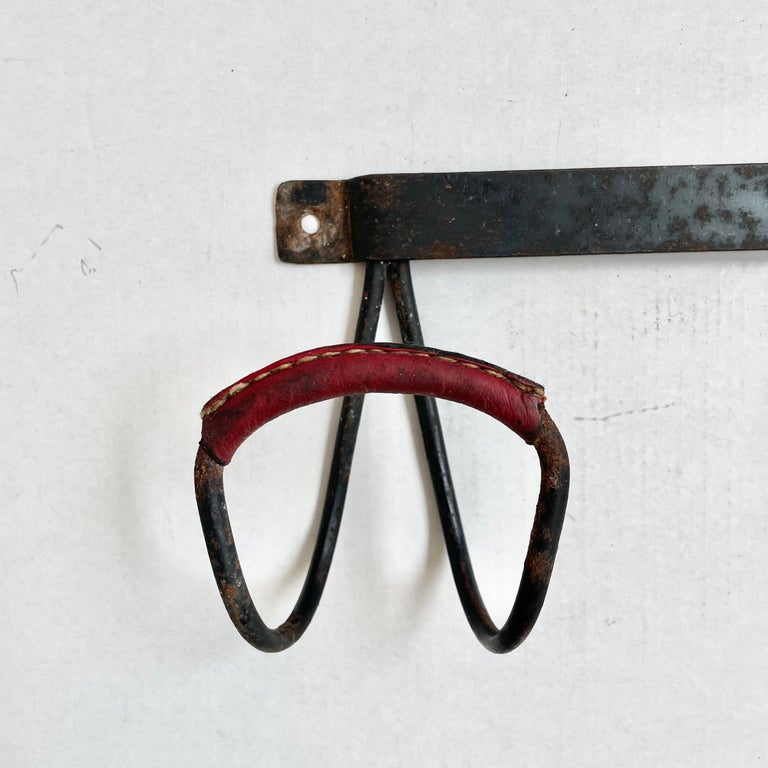 Handsome wall rack by French designer Jacques Adnet. Frame made of solid iron and features two large tongue hooks with red oxblood leather detailing on each hook. Signature Adnet contrast stitching. Good vintage condition. Two available. Priced