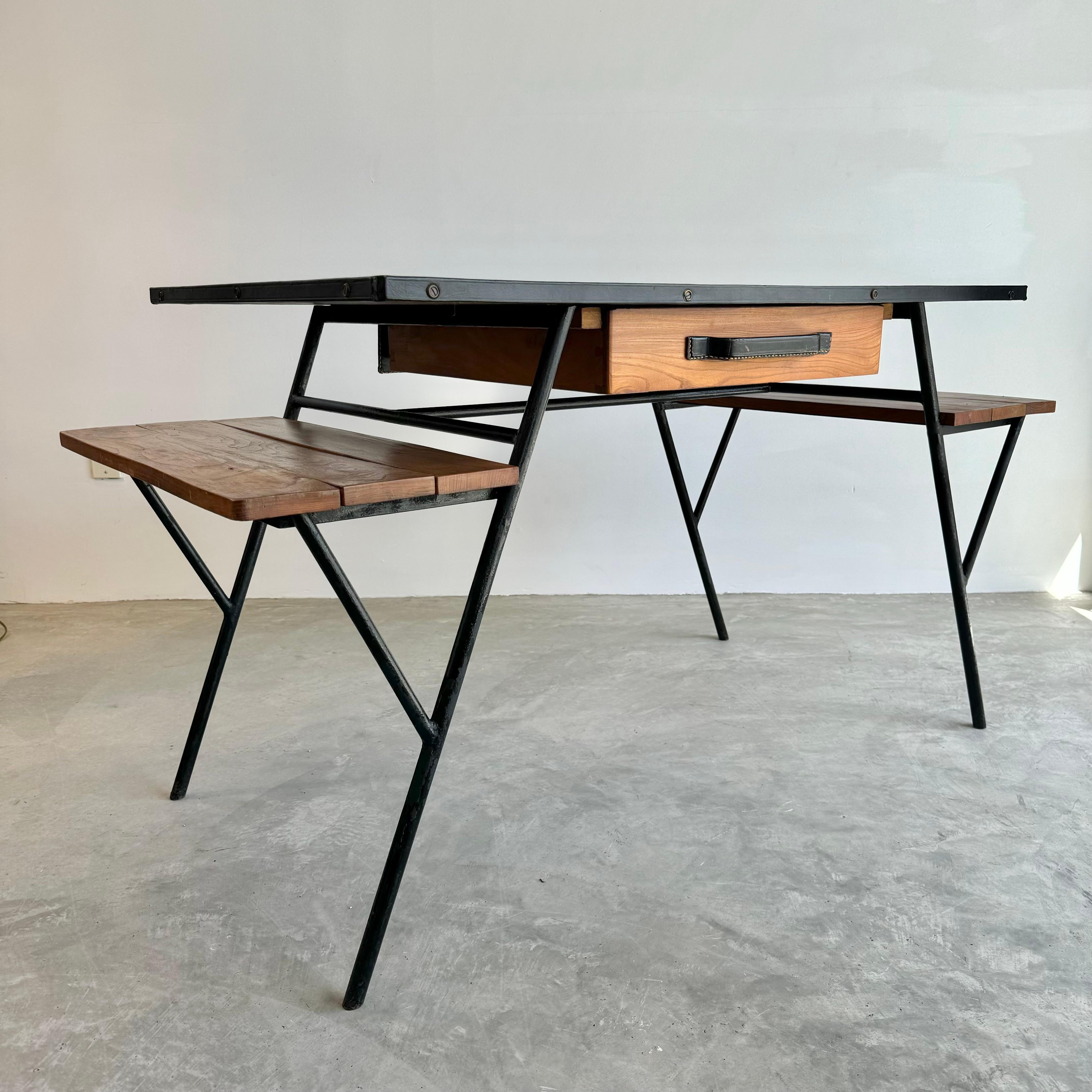Monumental Art Deco era desk by French Modernist designer Jacques Adnet. Black iron 'A' frame with 4 angled iron legs. Solid oak table top wrapped in black leather with brass studs provides an ample writing surface. Smaller shelves made of walnut