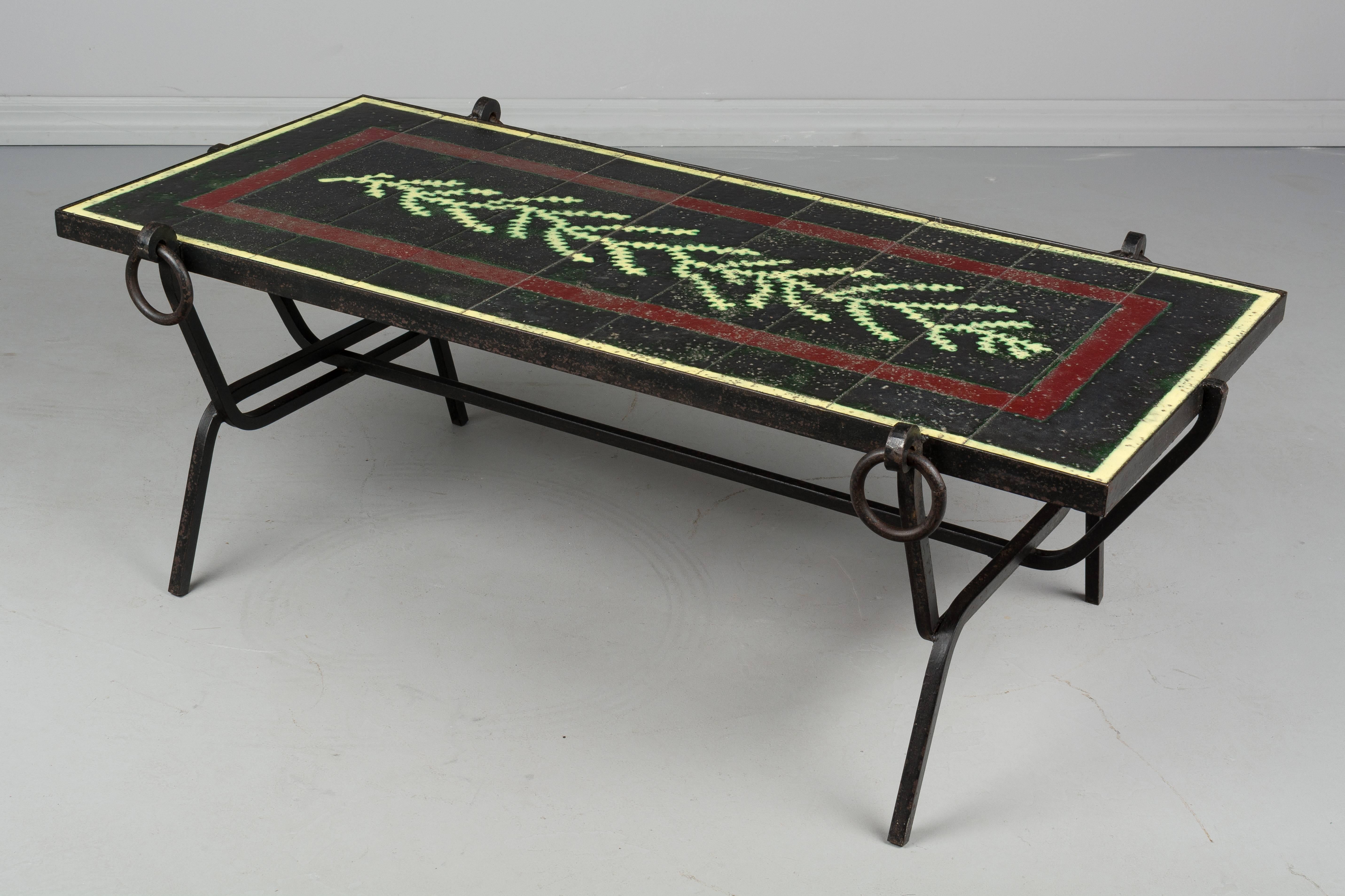 A Mid-Century Modern Jacques Adnet wrought iron table with glazed lava stone tile top. Nine tiles form a green vine or seaweed design on a black ground with burgundy border and yellow outline around the perimeter. Tiles are in excellent condition