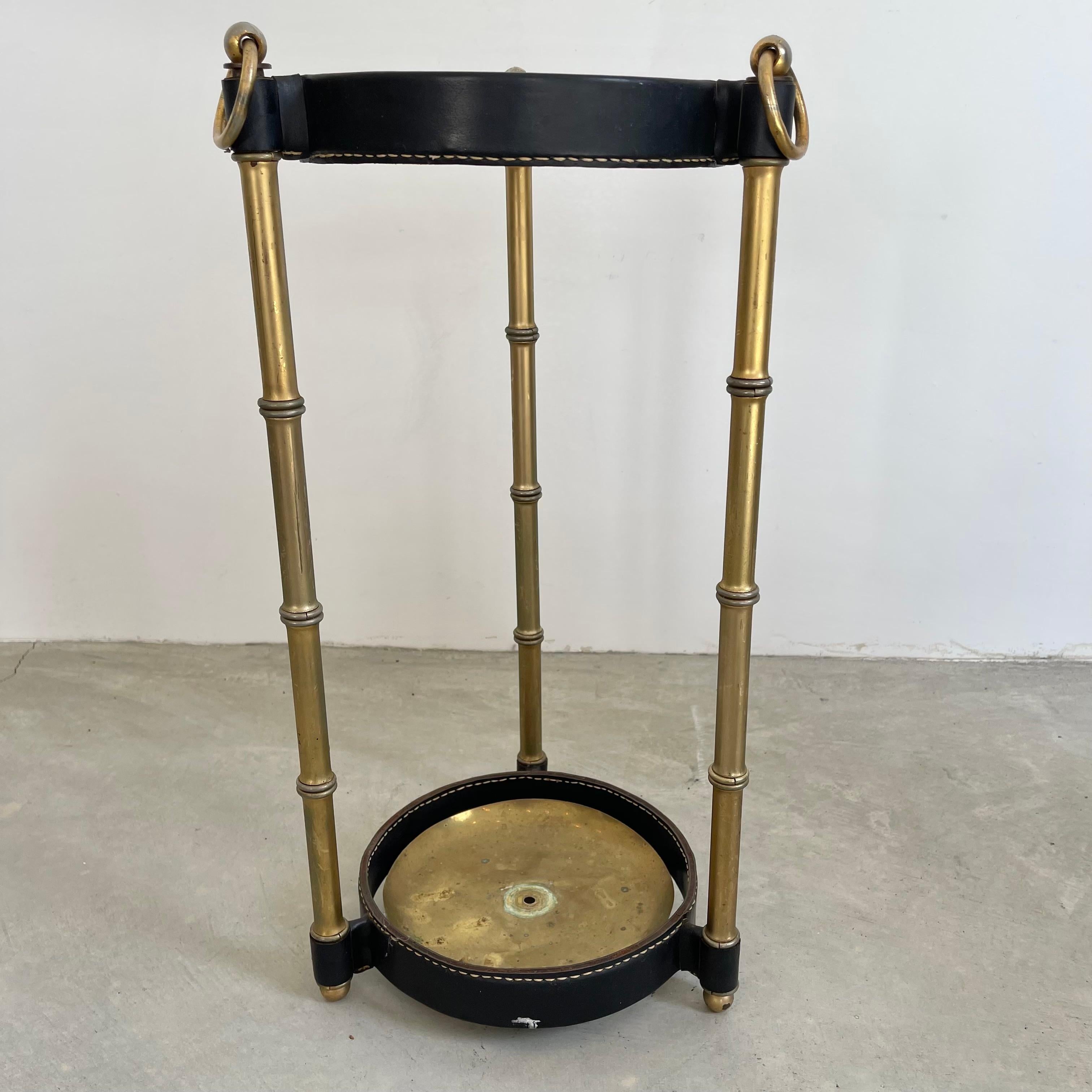 Handsome umbrella stand by Jacques Adnet. Metal upper and lower rings of the frame are wrapped in black leather stitched together with Adnet's signature contrast stitching. Three brass bamboo legs make up the frame with a brass dish at the bottom to