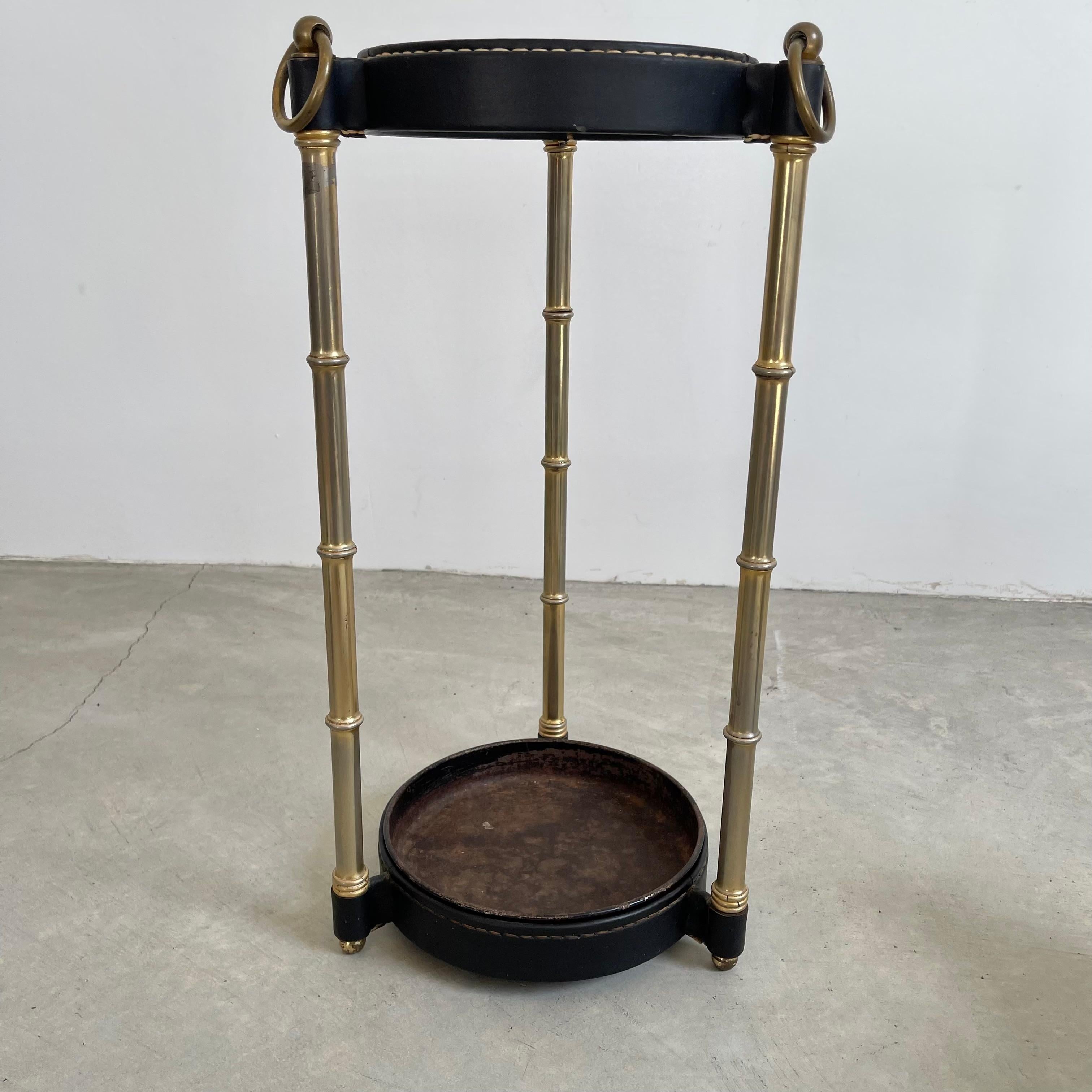 Handsome umbrella stand by Jacques Adnet. Metal upper and lower rings of the frame are wrapped in black leather stitched together with Adnet's signature contrast stitching. Three brass bamboo legs make up the frame with a metal dish at the bottom to