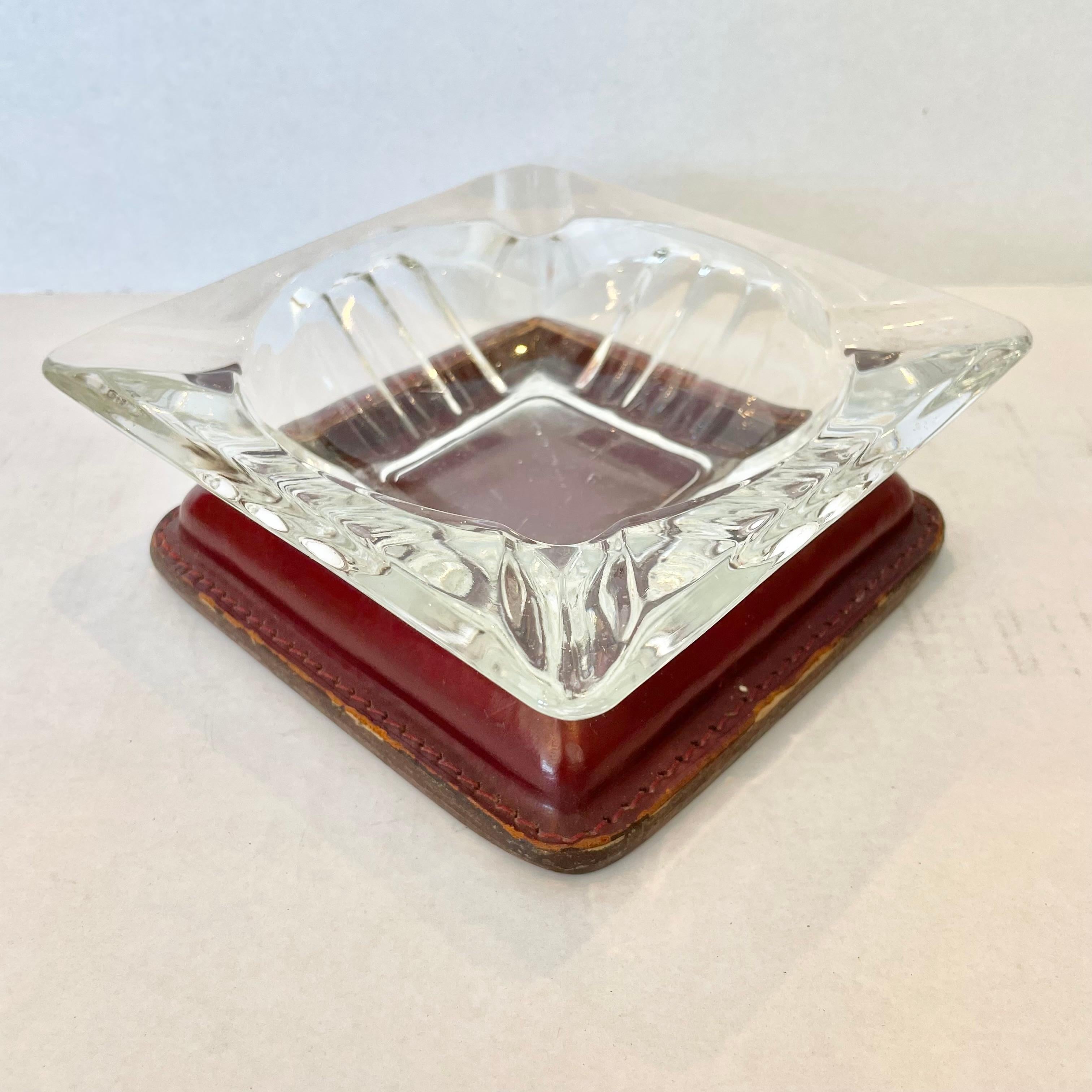 Classic leather and glass catchall / ashtray by French modernist designer Jacques Adnet. Rich oxblood leather base with stitching around the edges and a circular inset cutout. Crystal glass with circular base sits nicely inside with a cigarette