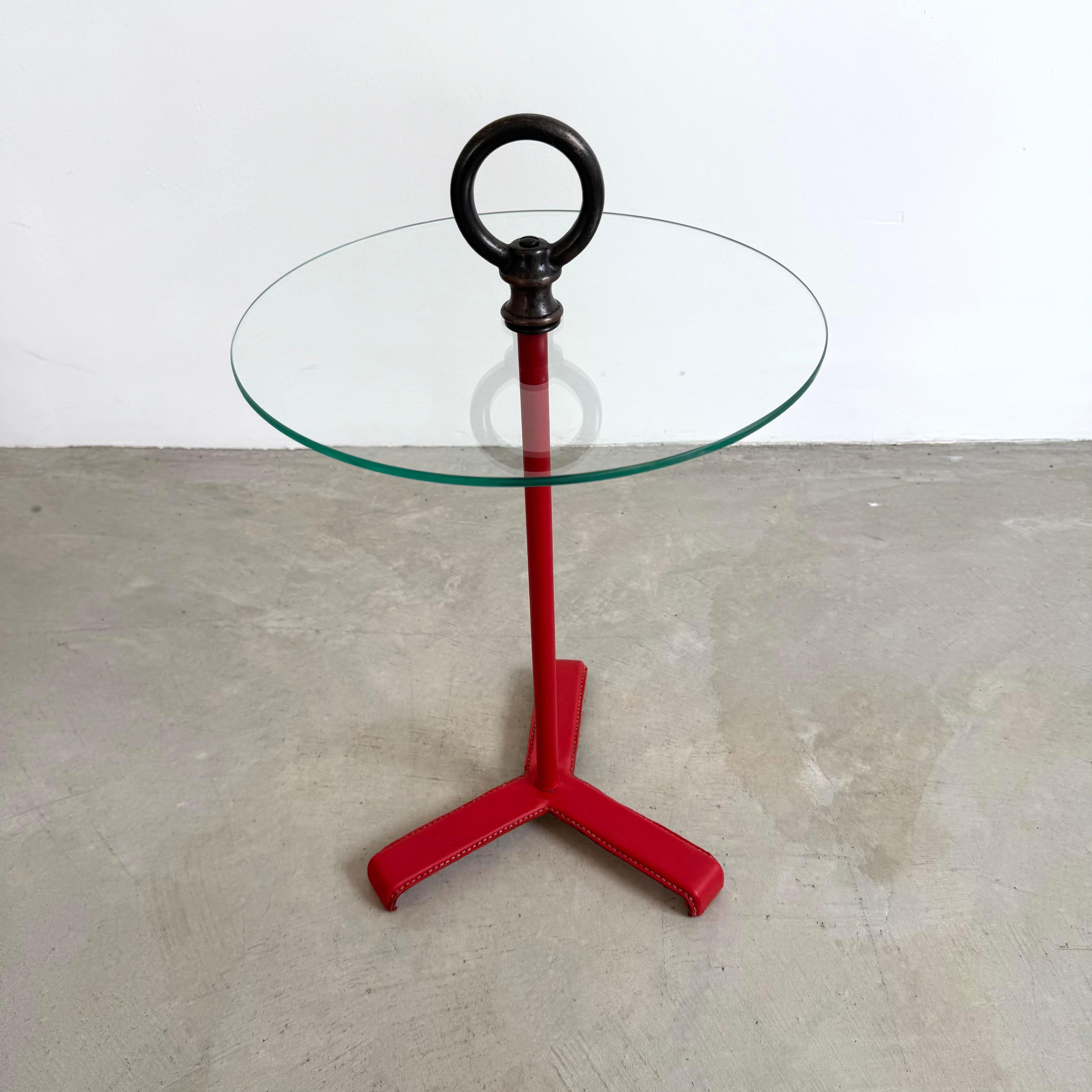 Tables d'appoint surdimensionnées en cuir et verre de Jacques Adnet, fabriquées en France dans les années 1950. Cuir rouge classique avec surpiqûres contrastées. Grande boucle en laiton antique sur le dessus. Deux disponibles. Les prix sont fixés à