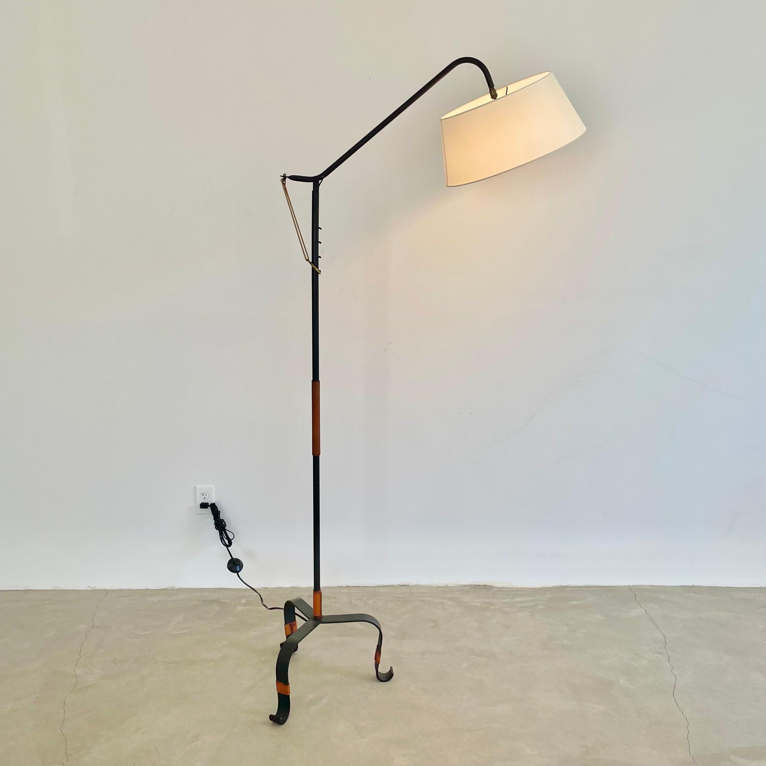 Handsome iron and leather floor lamp by French designer Jacques Adnet. Circa 1950s. Saddle leather trim on the feet and stem of the lamp. Saddle leather strap extending from the adjustable arm. This lamp has four arm heights adjustable by iron pegs