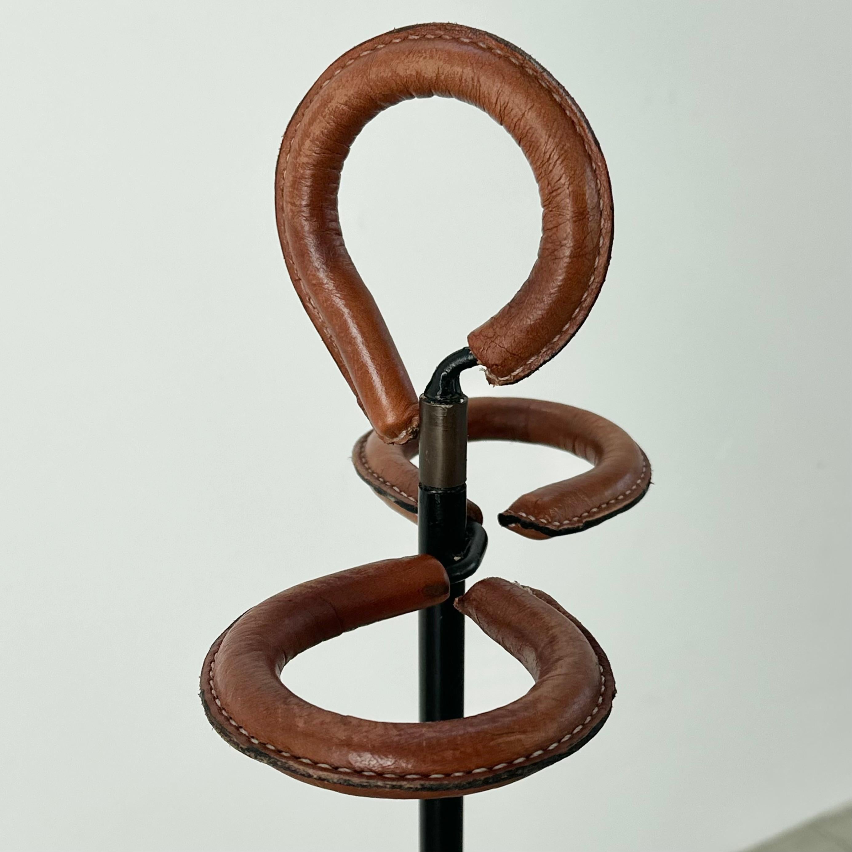 Absolutely stunning umbrella stand designed by Jacques Adnet in 1950s France. Sleek black iron tripod base creates a strong foundation for this classic piece. A caramel brown leather coats the three rungs that adorn the top of the