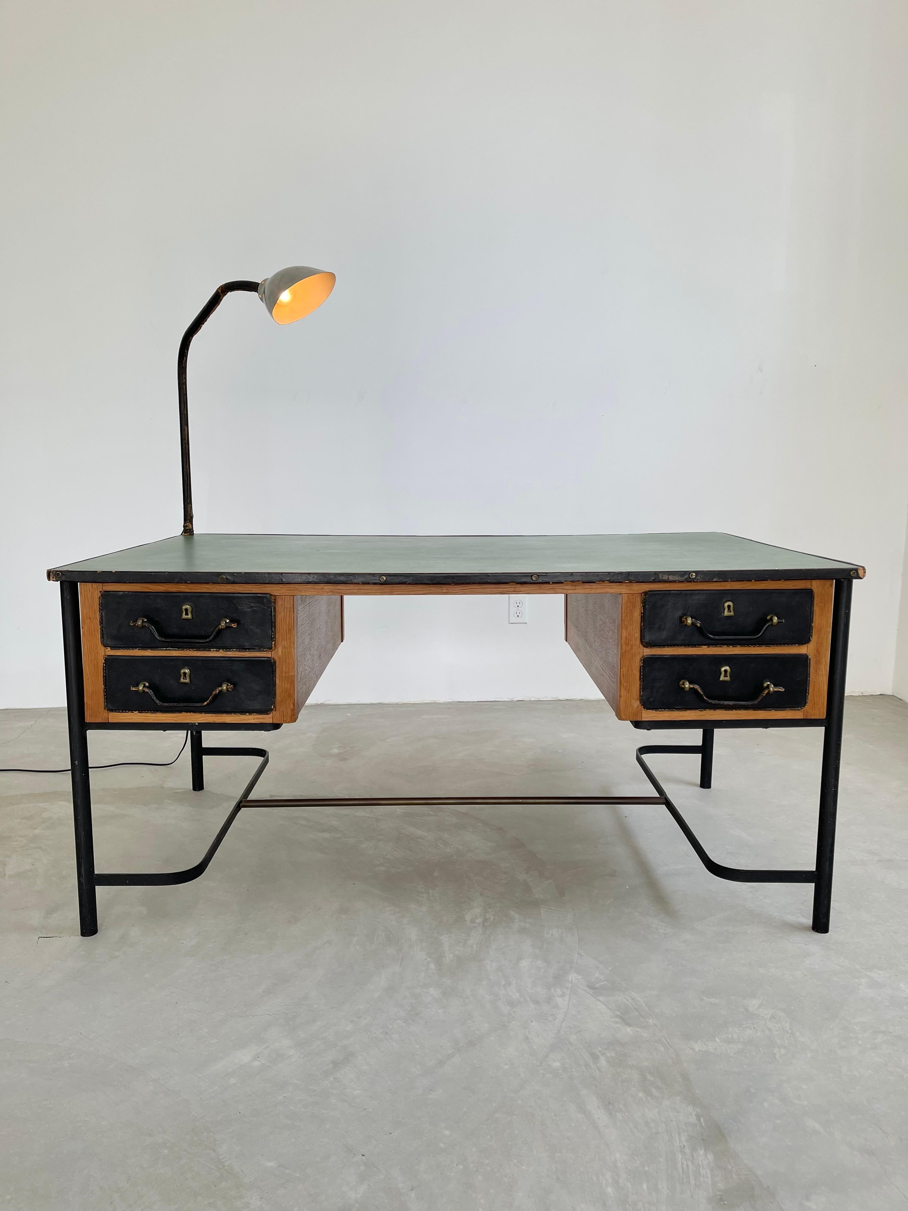 Monumental Art Deco era desk by French Art Deco Modernist designer Jacques Adnet. Iron frame with 4 posts, inverted u-shaped bases and brass support bar/foot rest connecting the two. Heavy oak frame wrapped in black leather with brass hardware. Two