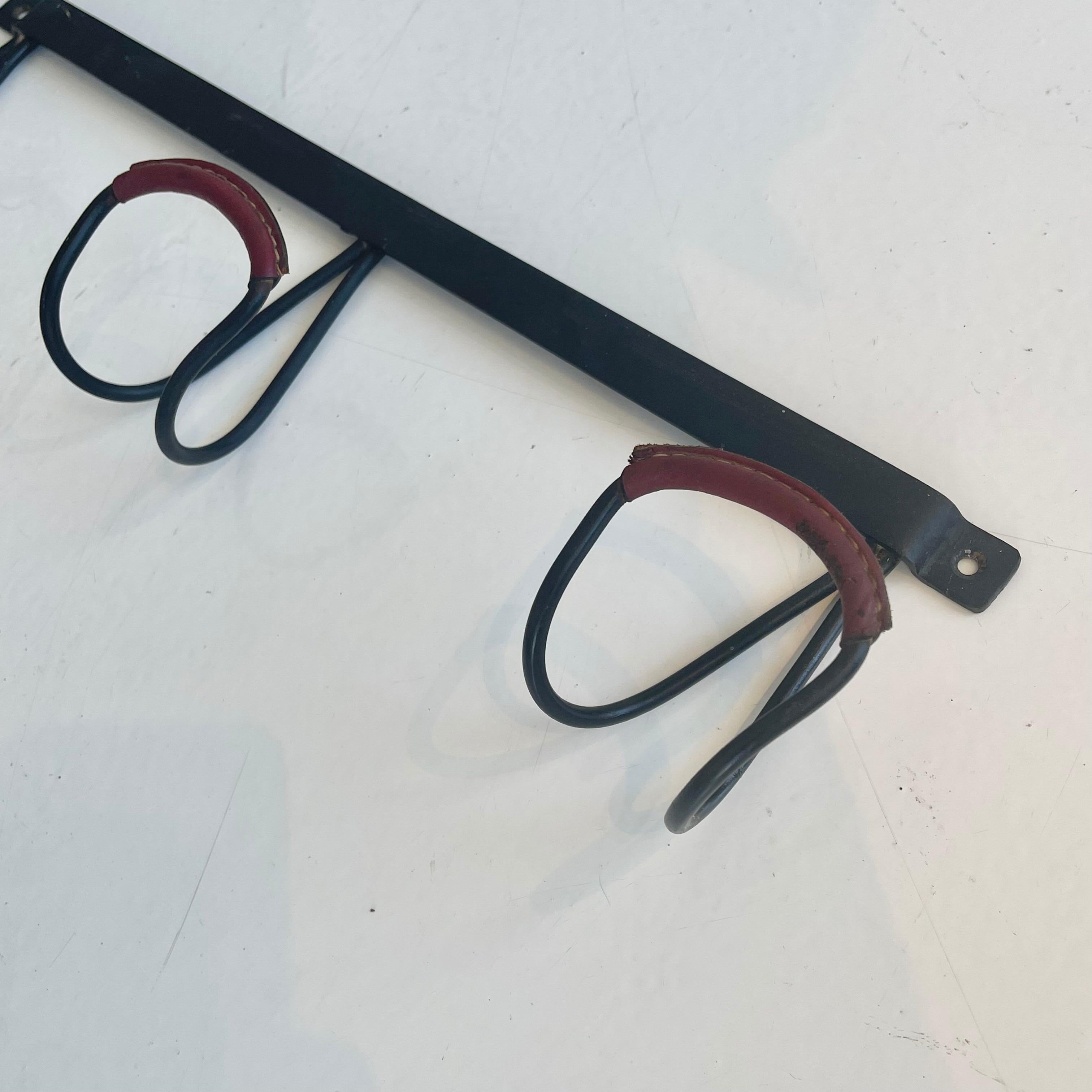 Handsome wall rack by French designer Jacques Adnet. Frame made of solid iron and features three large tongue hooks with red oxblood leather detailing on each hook. Signature Adnet contrast stitching. Good vintage condition. 

