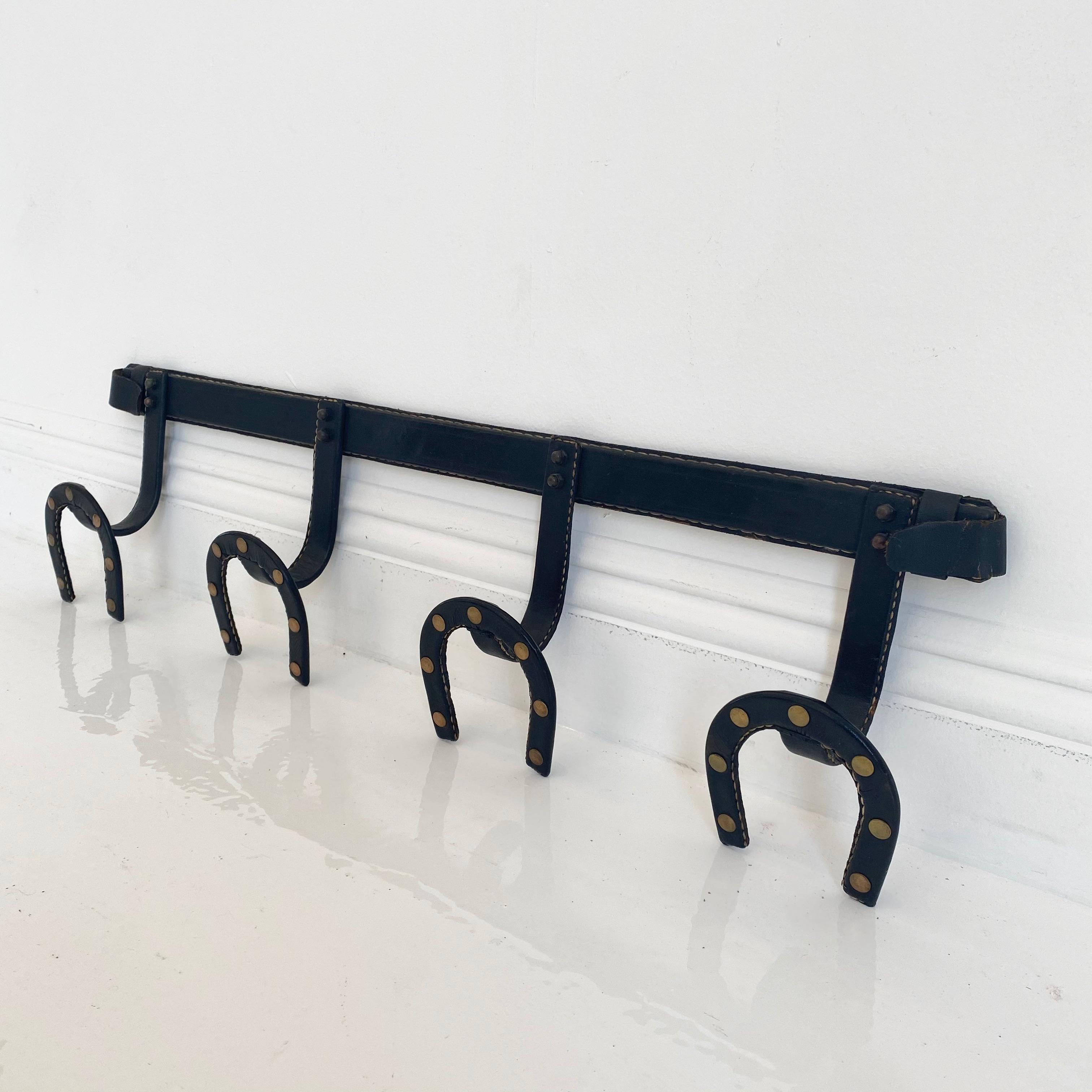 Handsome leather horse shoe coat rack by French designer Jacques Adnet. Good vintage condition. Iron frame completely wrapped in black saddle leather. 4 horseshoe shaped hooks with brass detailing. Fantastic piece of collectible Adnet.
