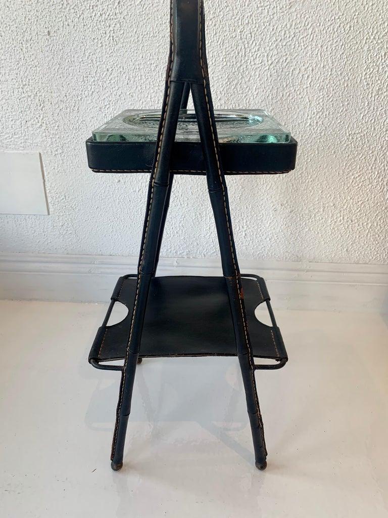 Stunning leather side table / catchall by Jacques Adnet. Iron A frame completely wrapped in black leather with 2 tiers. Glass tray on top and a leather tray underneath as well. Signature Adnet contrast stitching. Brass ball feet. Good vintage