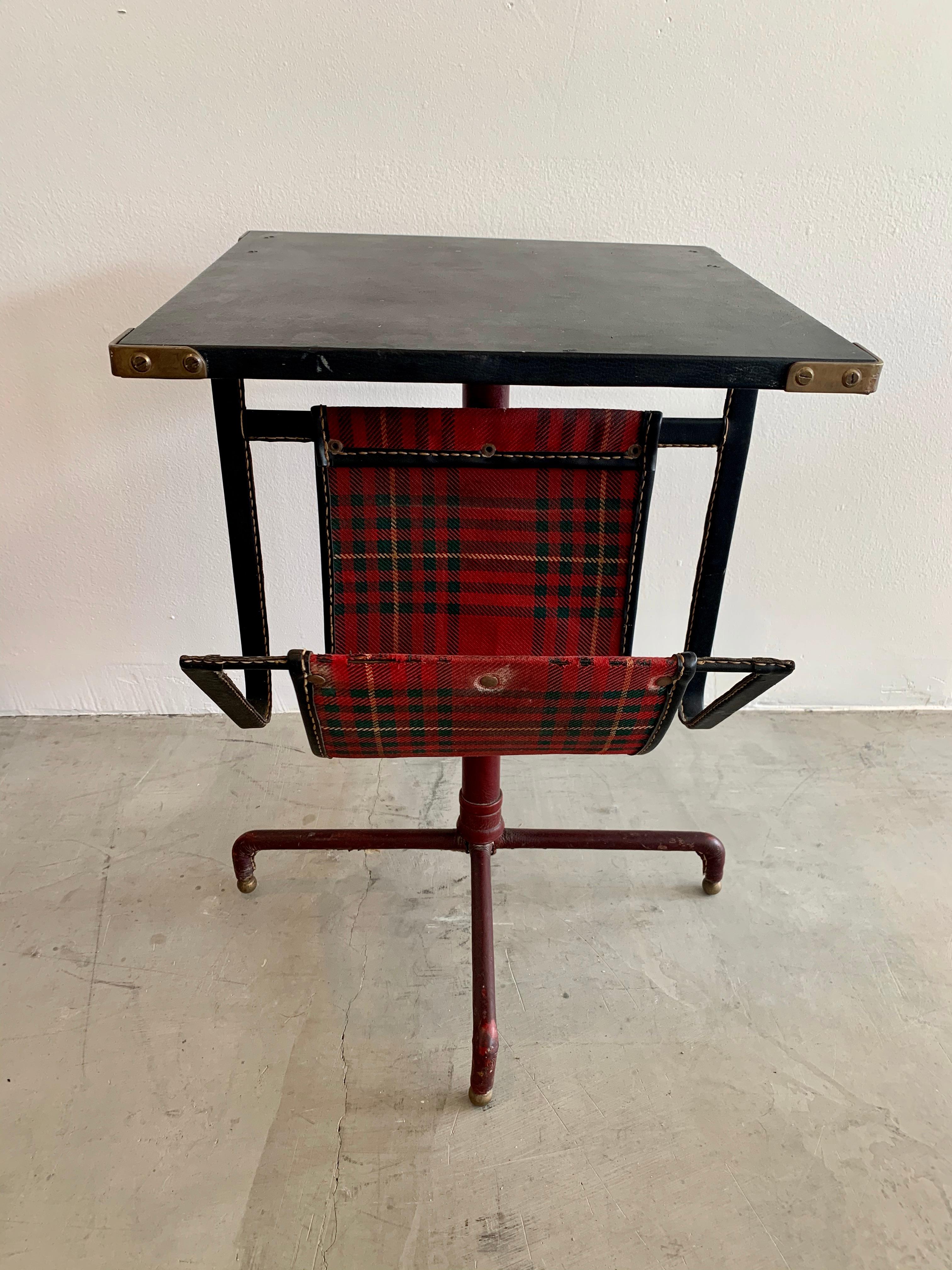 Very unusual side table by Jacques Adnet. Completely wrapped in leather. Tabletop is black leather with brass corners. Frame is also wrapped in black leather and extends down, holding a plaid and leather shelf for holding books or magazines. Base is
