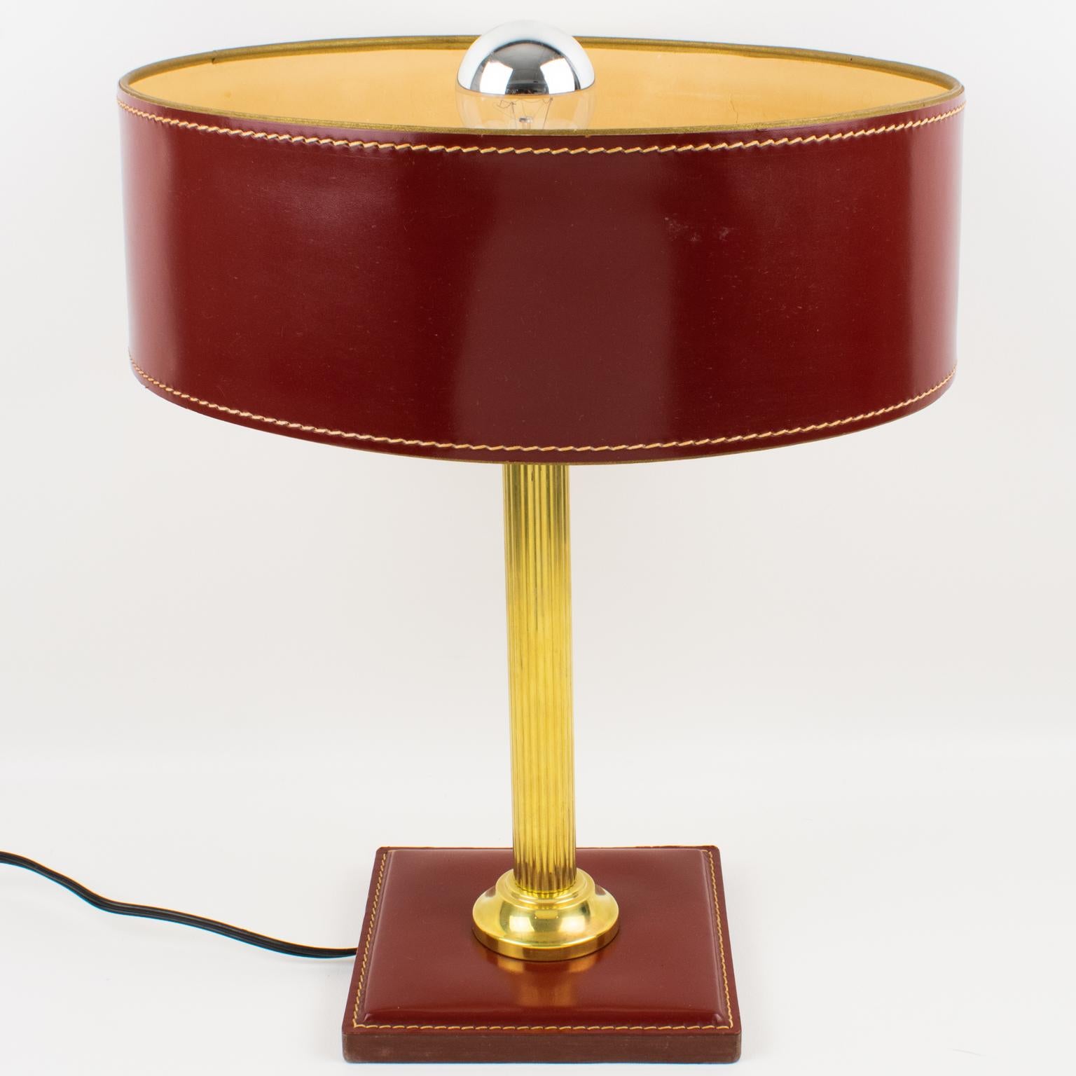 Elegant table lamp designed by Jacques Adnet (1901 - 1984). A classical burgundy-red color lamp features a hand-stitched shade with ivory paper lining, polished brass reeded column stem, and square base in the same hand-stitched burgundy-red