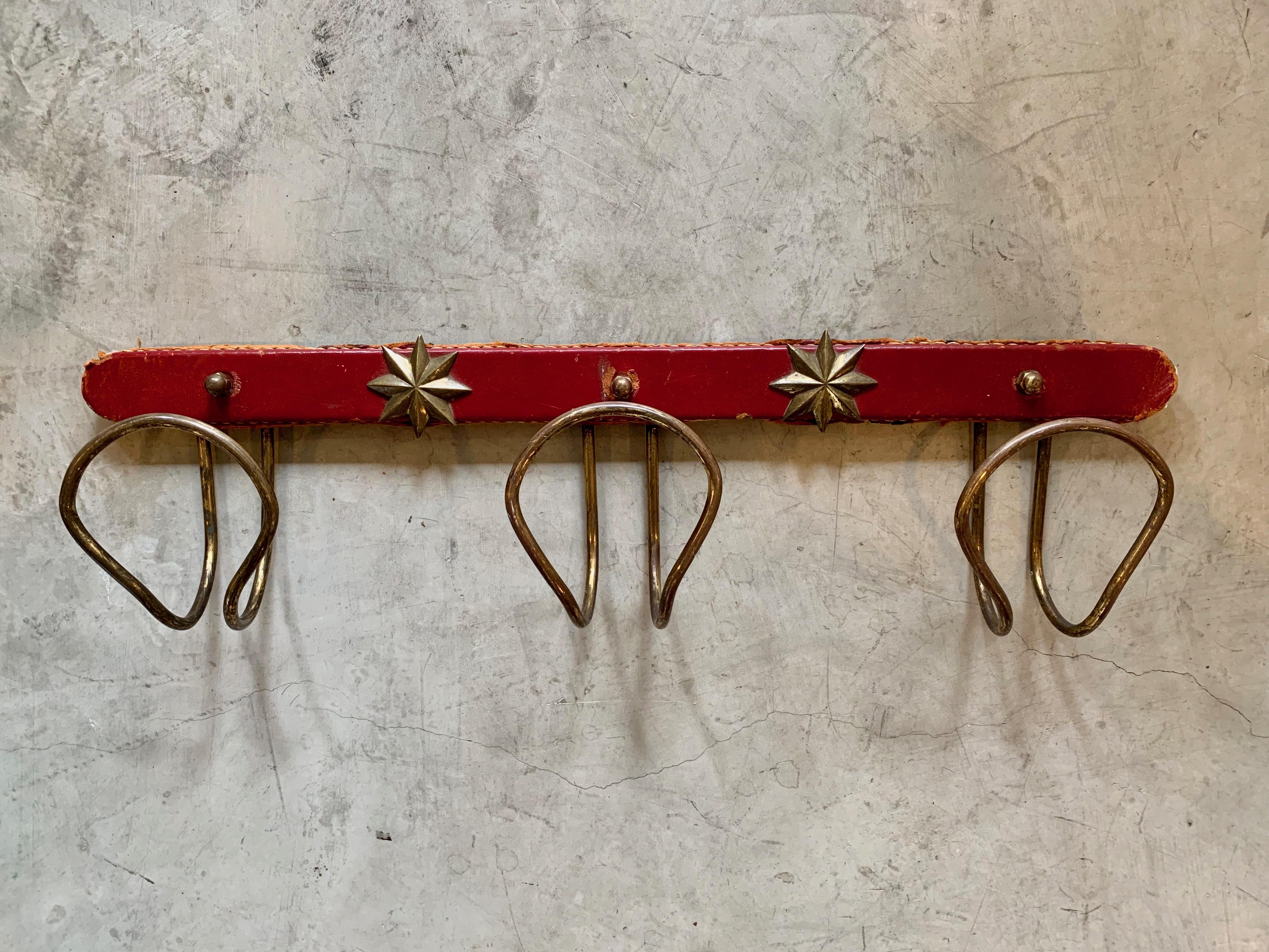 Classic coat rack by French designer Jacques Adnet. Three brass tongue shaped hooks extending from red leather bar with signature Adnet contrast stitching. Three brass nuts covering the wall mounting holes. 2 brass decorative stars. Leather in