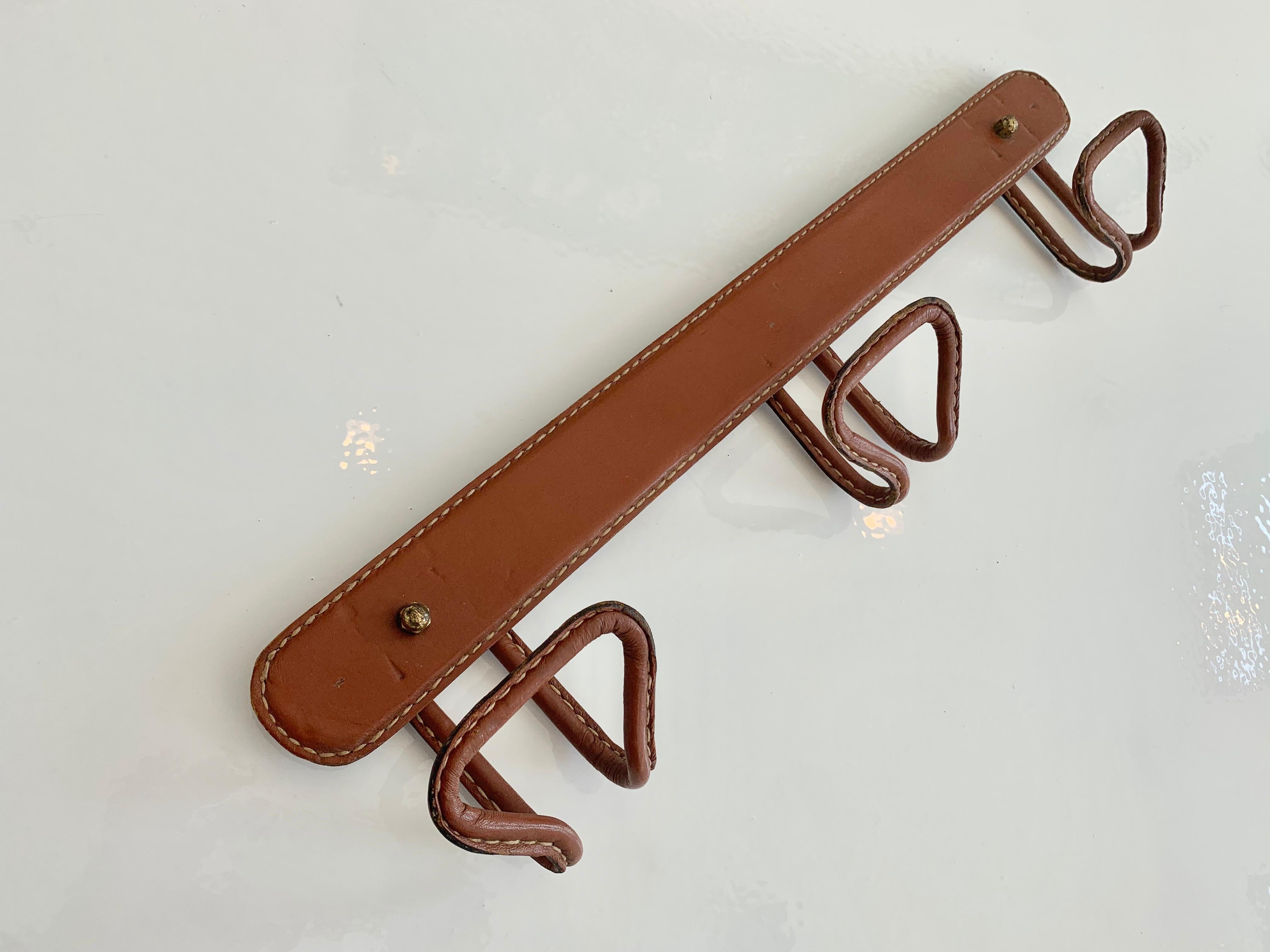 Elegant saddle leather coat rack by French designer Jacques Adnet. Iron frame with saddle leather tongue hooks. Entire piece is covered in leather. Two brass nuts enable you to hide the screws in the wall. Signature Adnet contrast stitching. Great