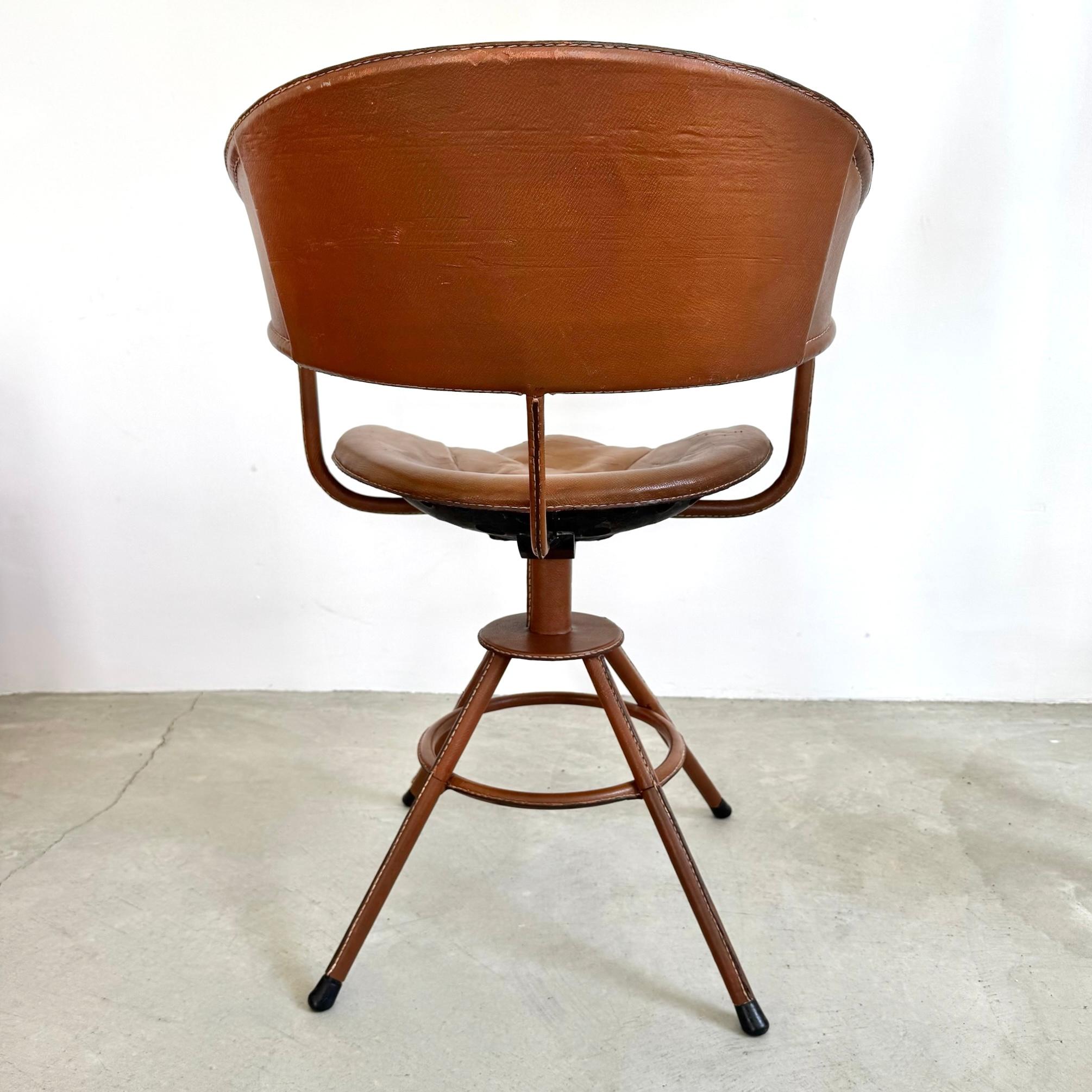 Lehnen Sie sich zurück auf diesem herrlichen Stuhl des französischen Designers Jacques Adnet. Ein Eisenrahmen ist komplett mit schönem, kamelfarbenem Leder umhüllt. Vier Beine bilden eine dreibeinige Basis. Die Rückenlehne ist unter dem